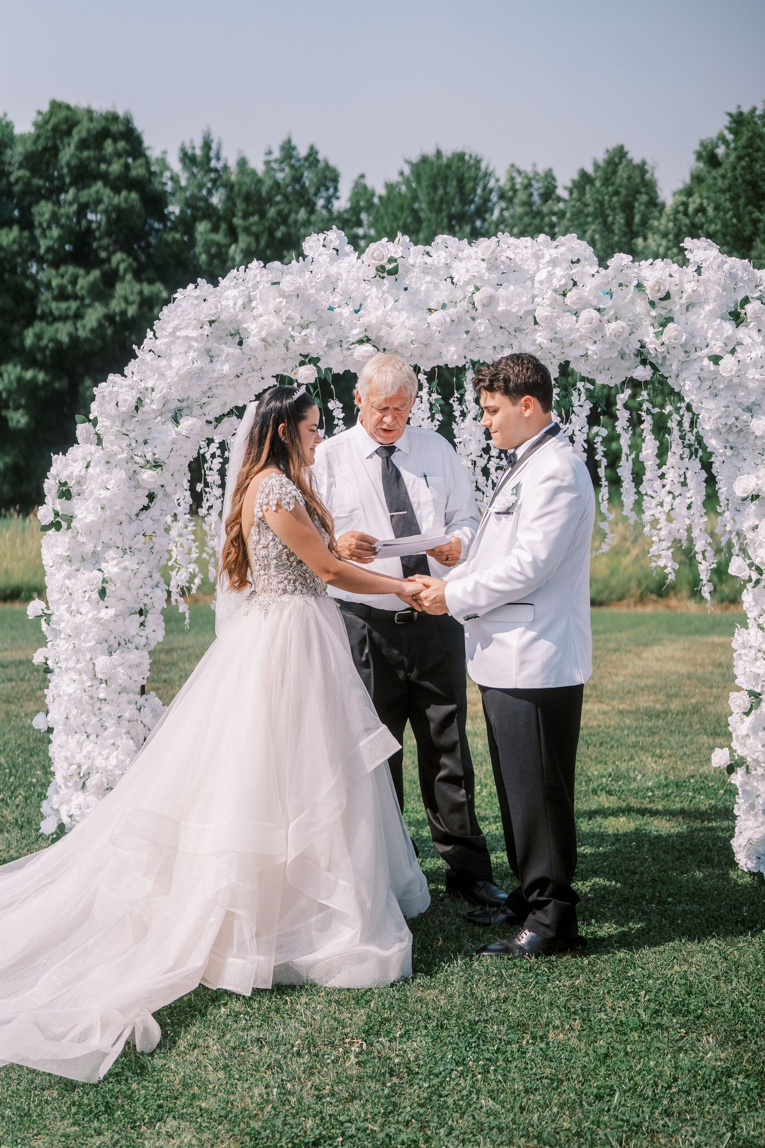 A wedding ceremony captured in sequence under a white floral arch on a bright outdoor lawn, showcasing candid moments.