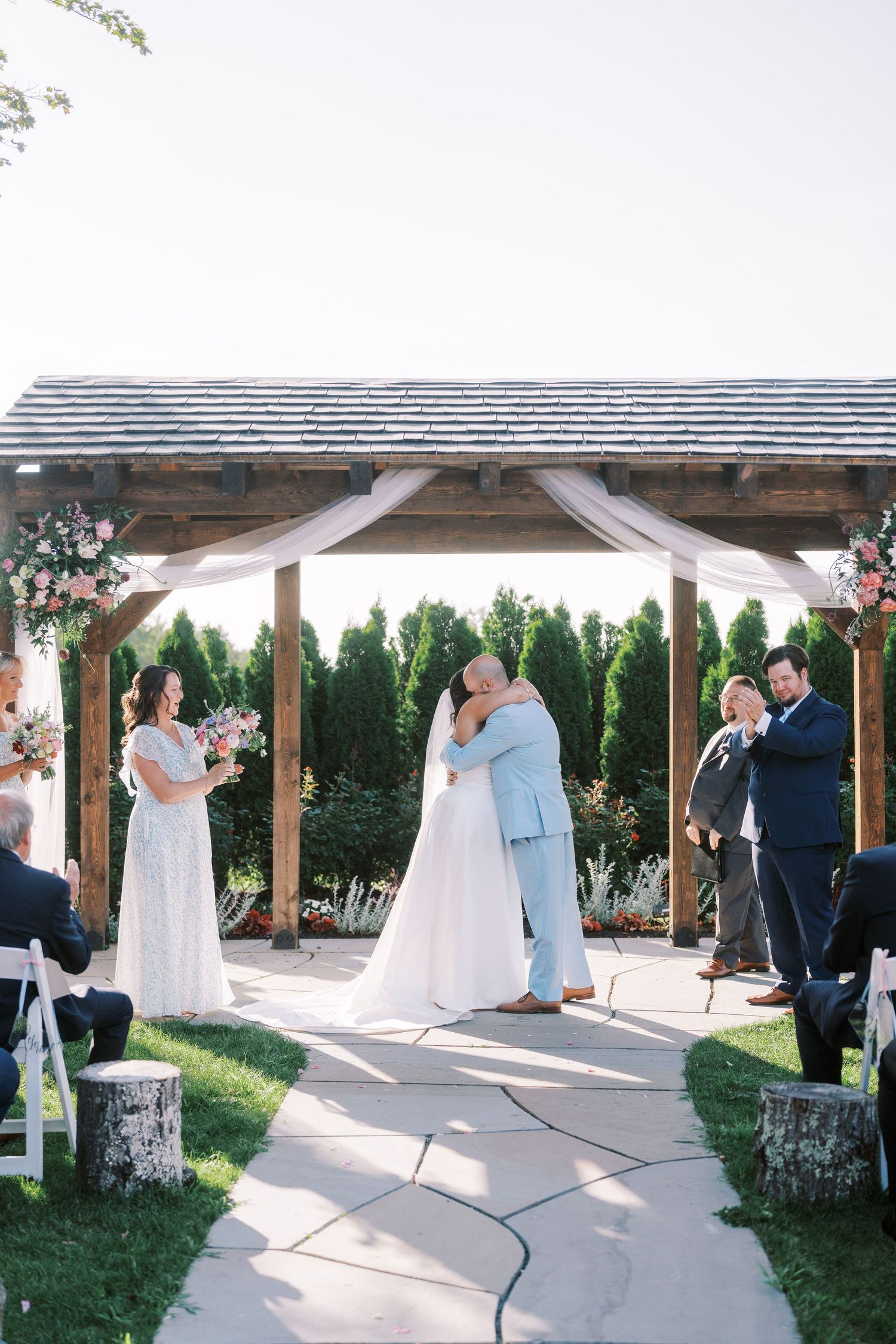Outdoor wedding ceremony under a rustic wooden pergola decorated with flowing fabric and flowers.