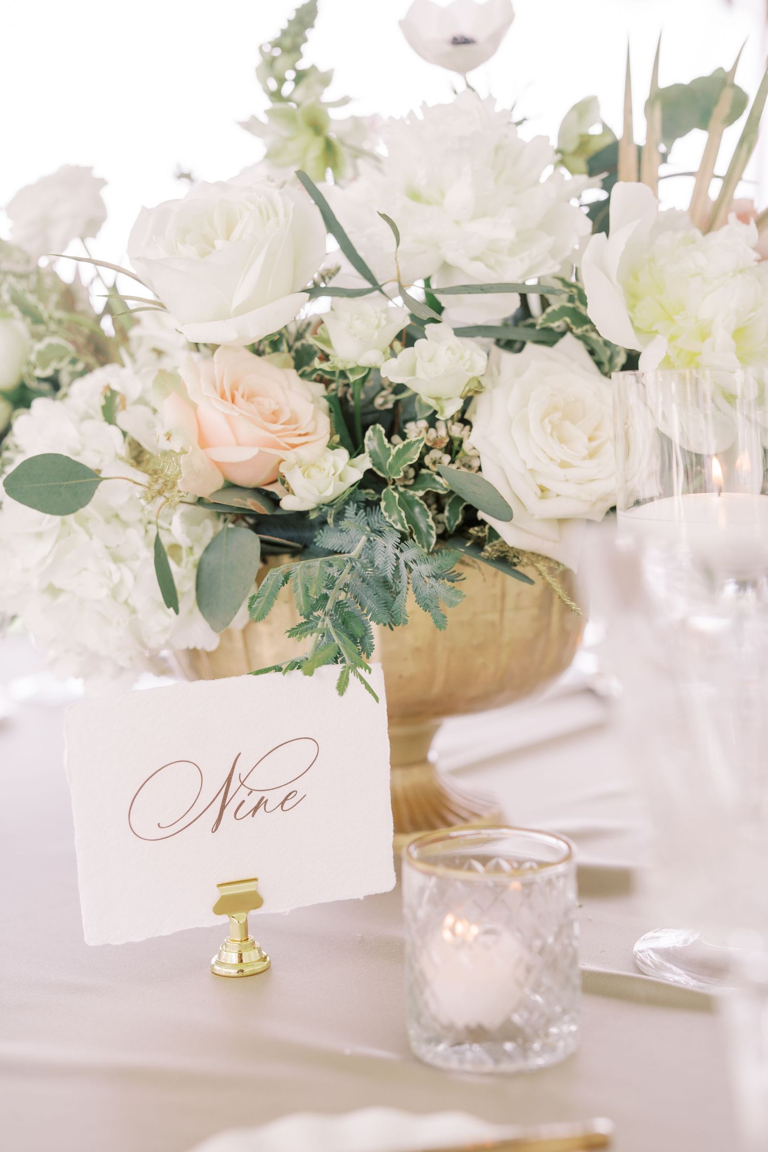 Elegant white floral table centerpiece with name cards and votives on crisp white linen for a luxurious wedding reception.
