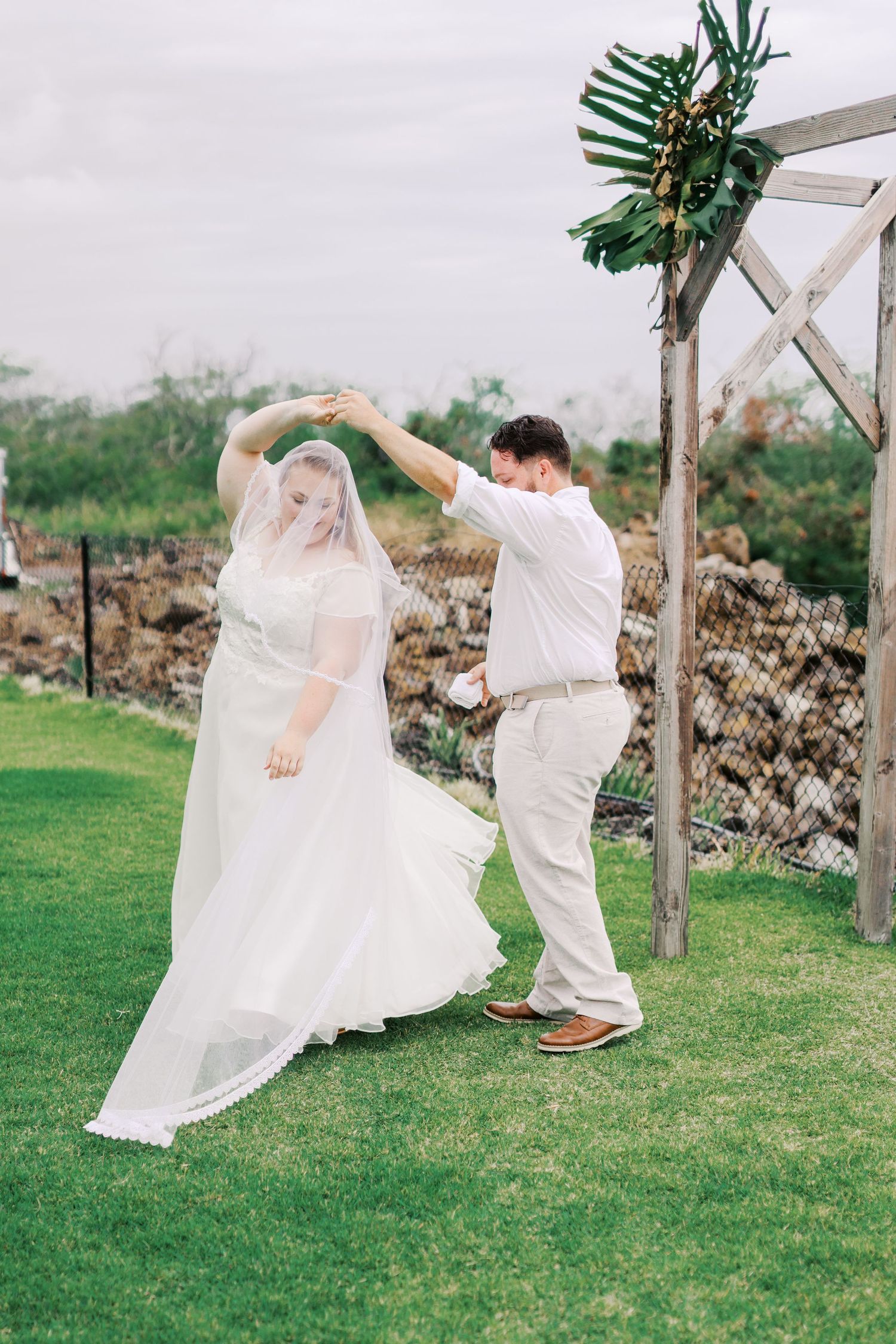 A romantic twirl on a grassy lawn beneath a wooden arch decorated with tropical greenery.