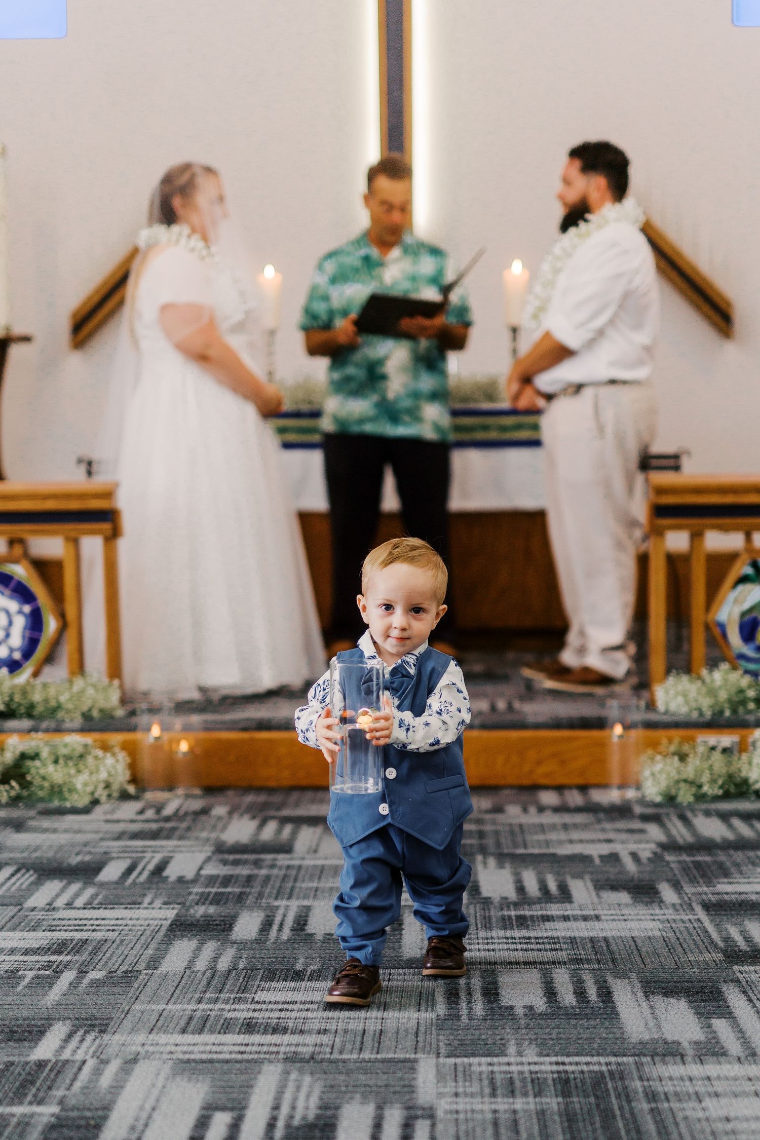 A toddler in blue overalls stands in front of adults during a wedding ceremony.