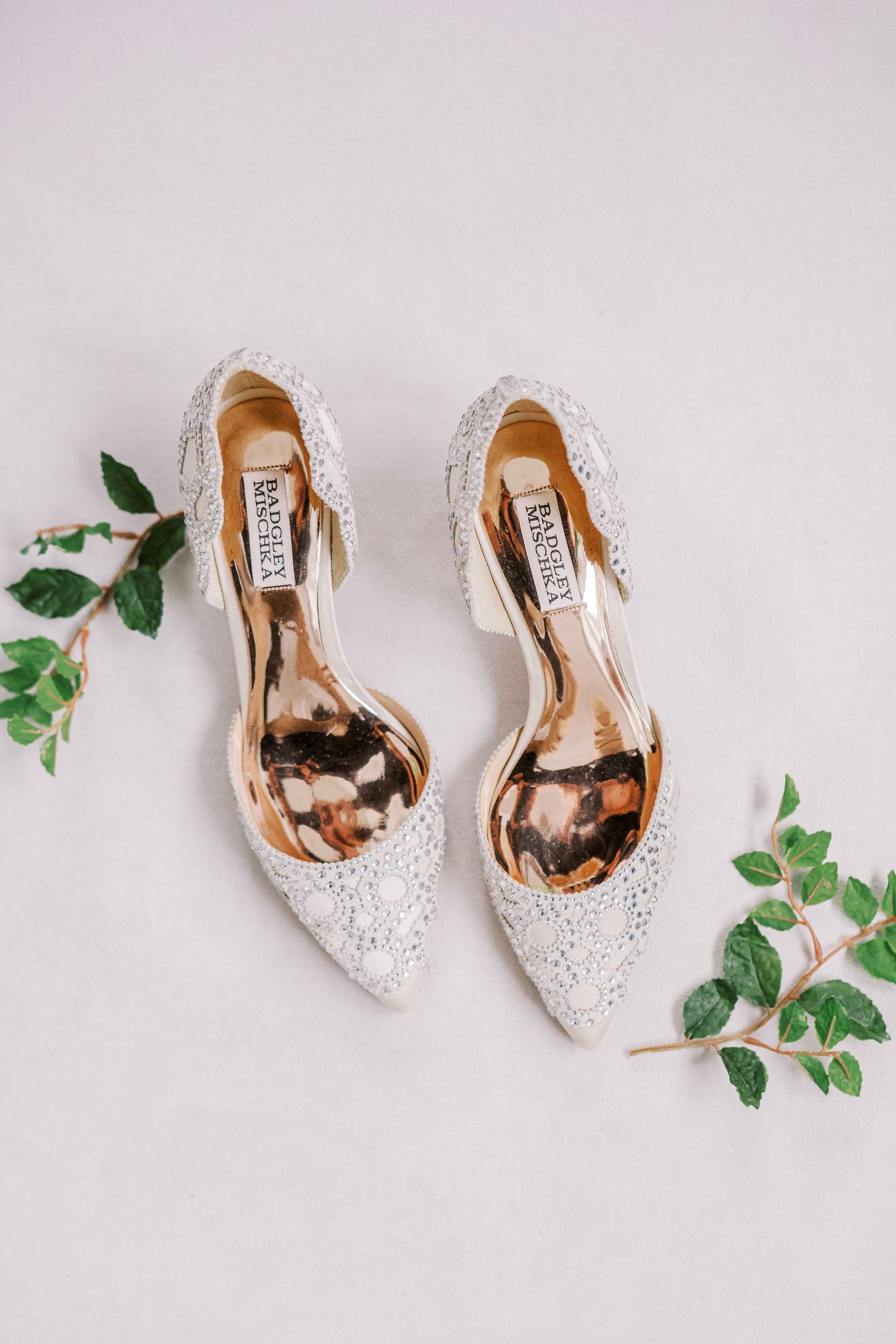 White lace Badgley Mischka bridal shoes with crystal details lie flat beside green ivy leaves on light background.