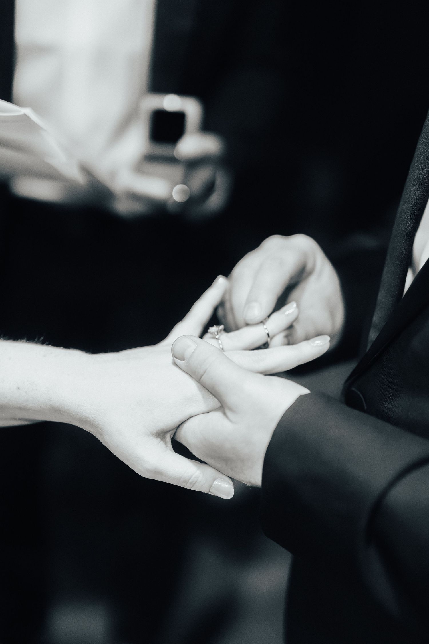 Close up photo of hands exchanging wedding rings during a ceremony against a dark background.