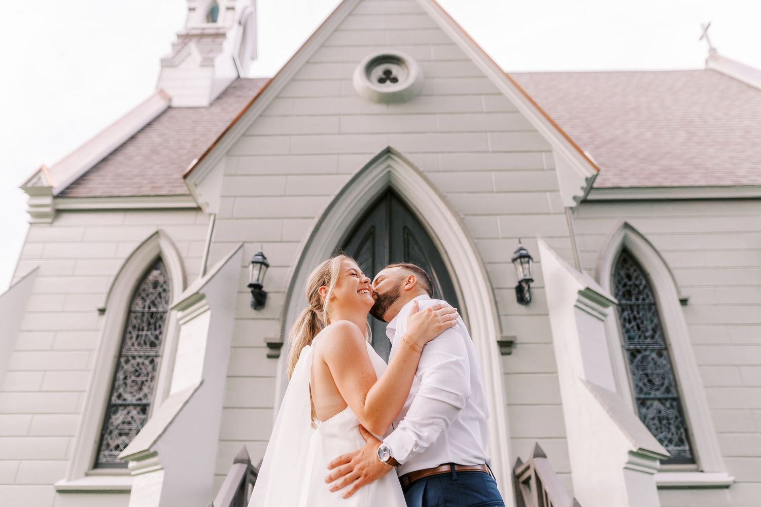 A bride and groom share an intimate embrace outside a white chapel with a series of decorative arched windows.
