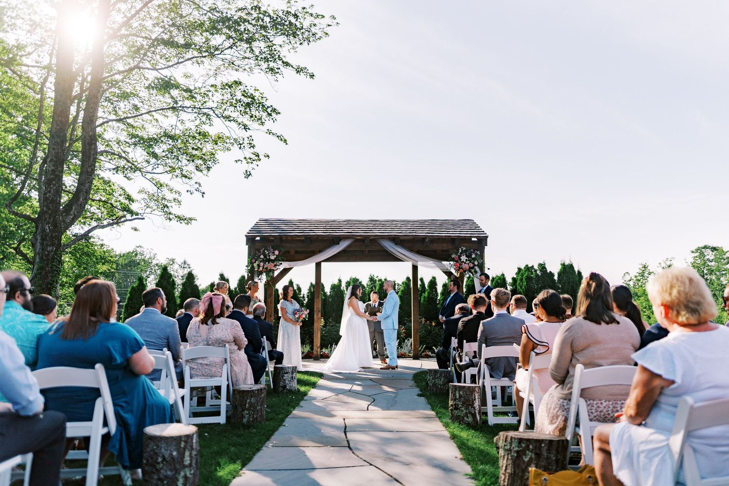 Outdoor wedding ceremony under wooden pavilion with guests seated on white chairs.