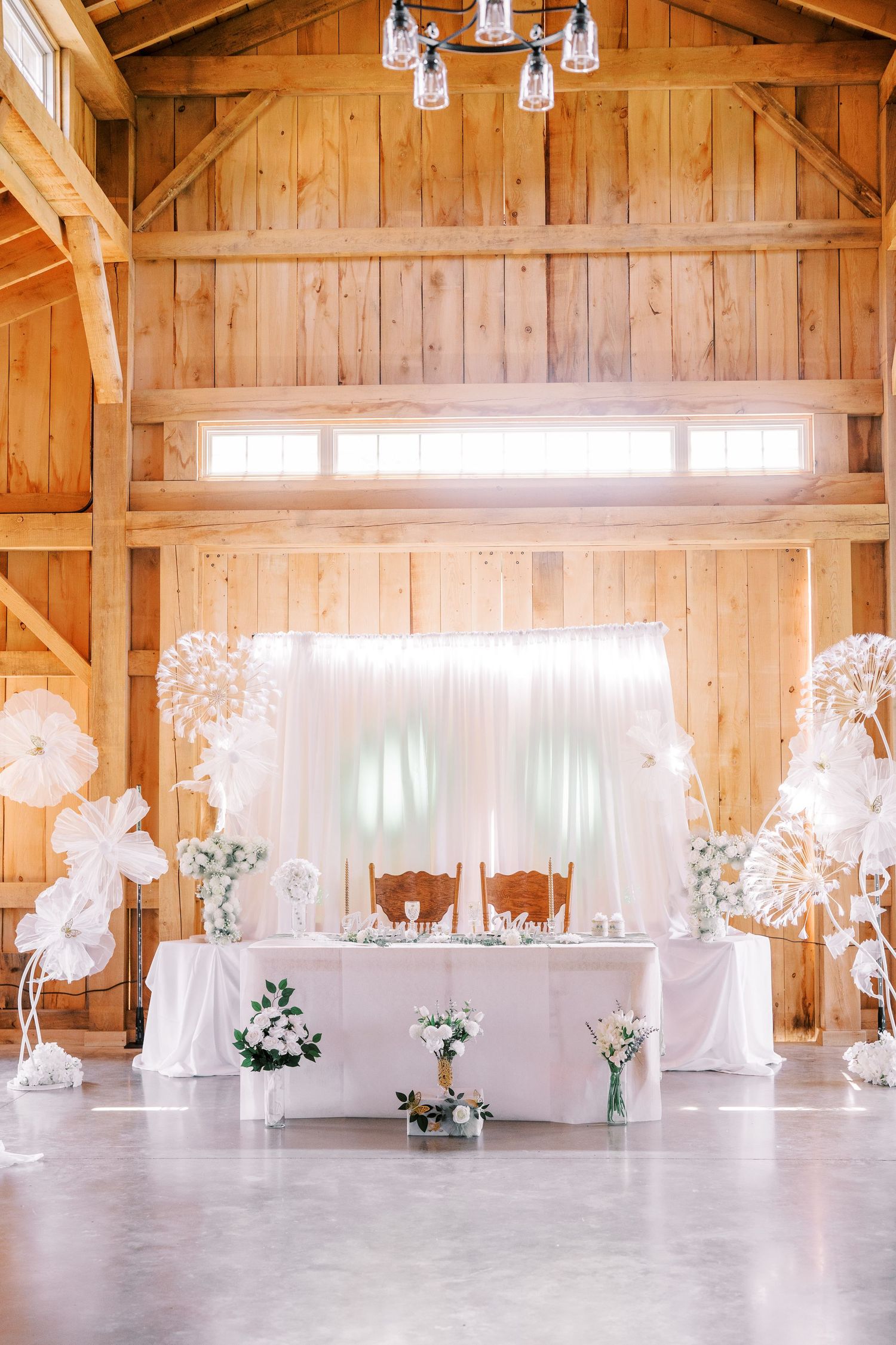 Elegant rustic barn wedding reception setup with white drapes, floral arrangements and a head table against wooden walls.