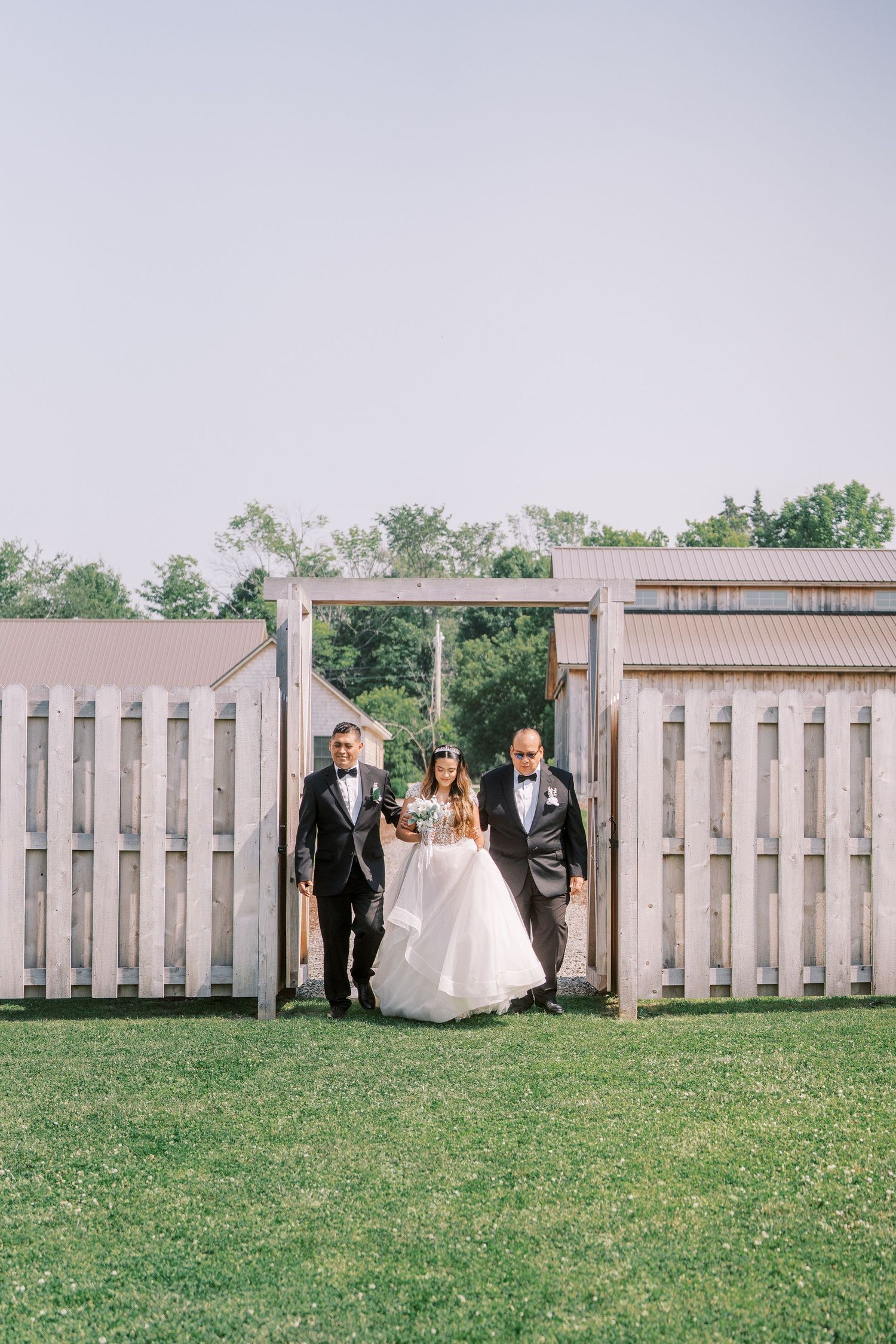 Three people in formal wedding attire walk through a rustic wooden gate on a grassy lawn at an outdoor venue.
