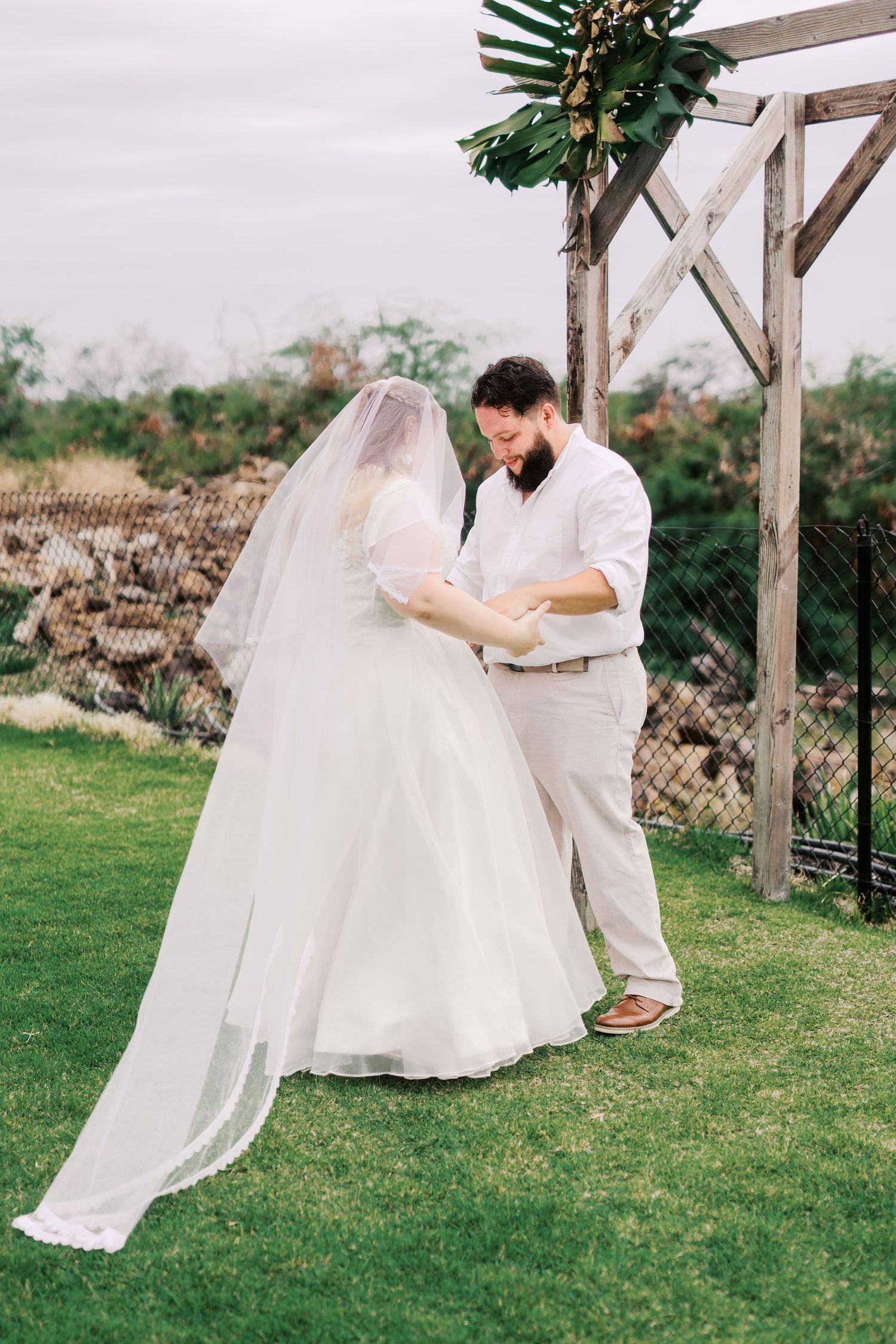 A couple embraces under a wooden wedding arch decorated with tropical greenery.