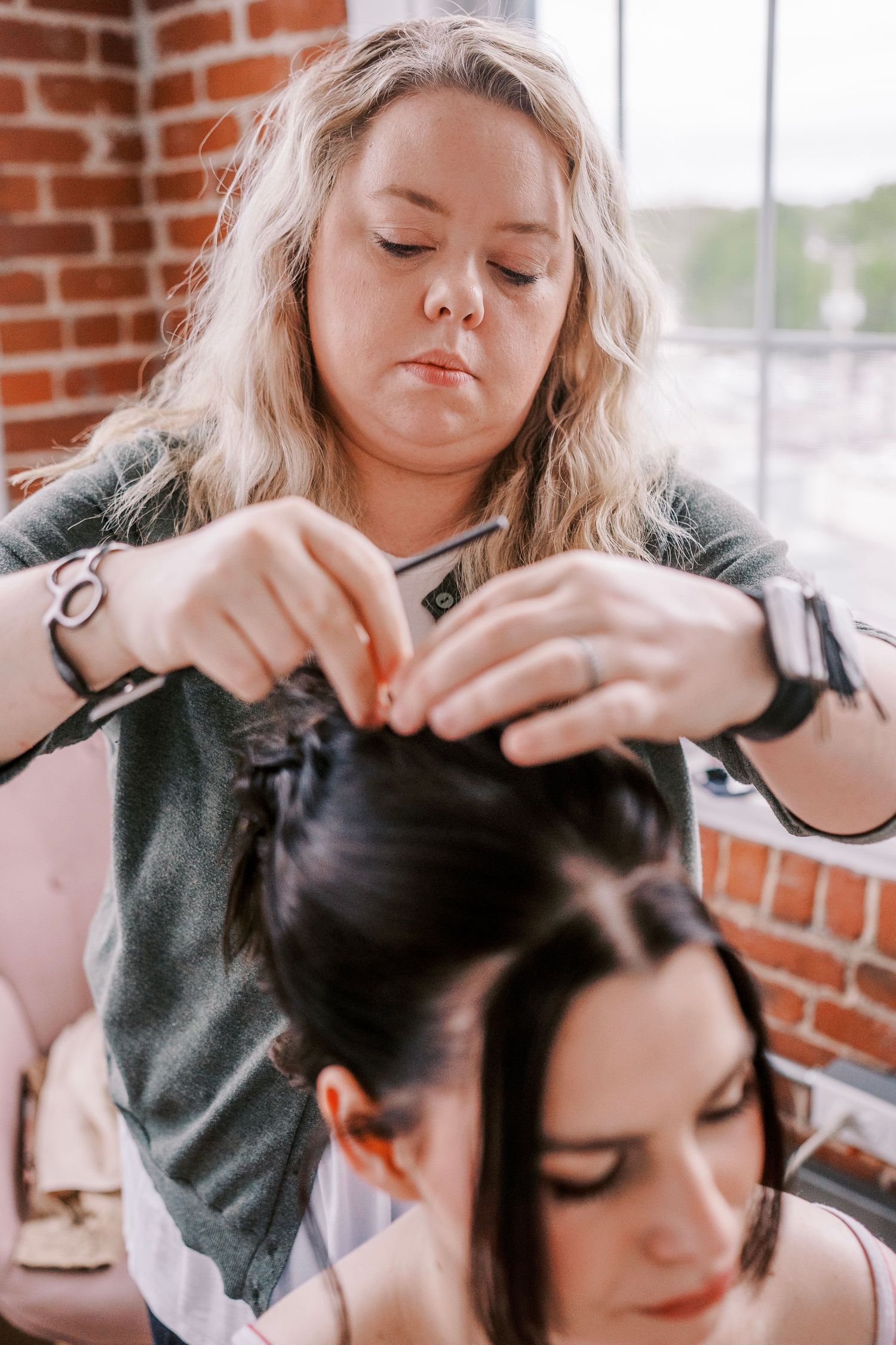 A stylist works on pinning up dark hair in a bridal preparation setting with exposed brick walls.