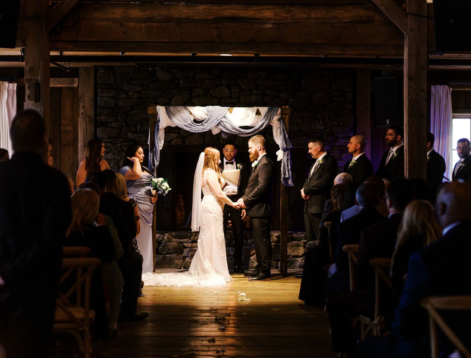 A couple stands at the altar during their rustic barn wedding ceremony illuminated by dramatic lighting.