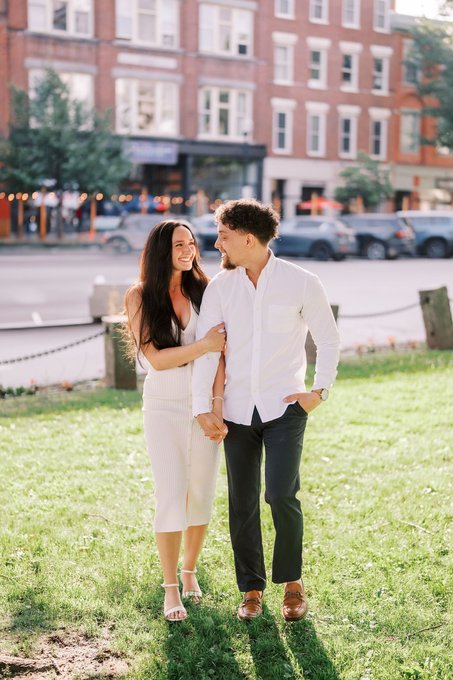 A stylish couple shares a tender moment on grass in front of a classic brick building in the city.