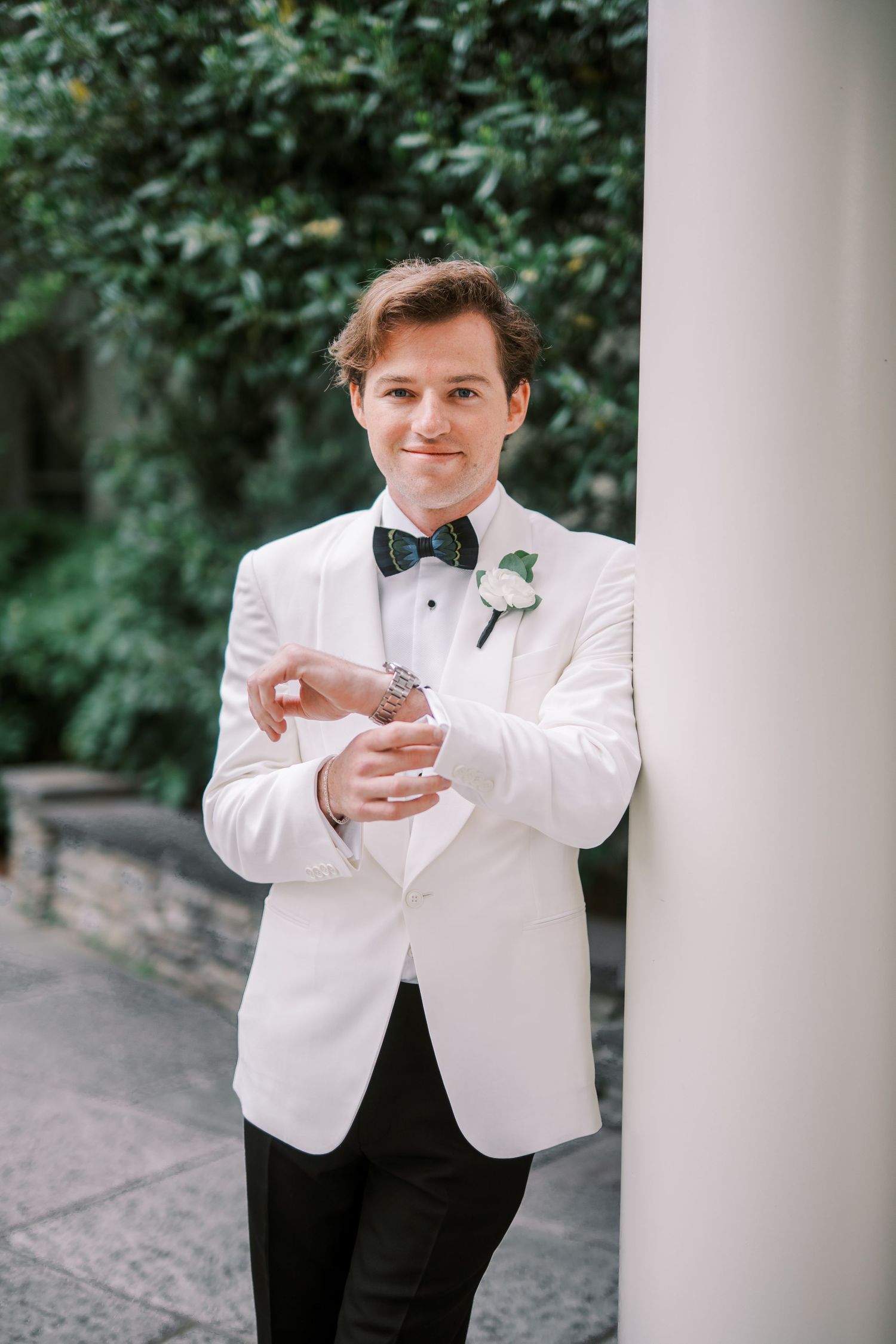 Well-dressed groom in classic white tuxedo jacket and black bow tie leans against elegant white column outdoors.
