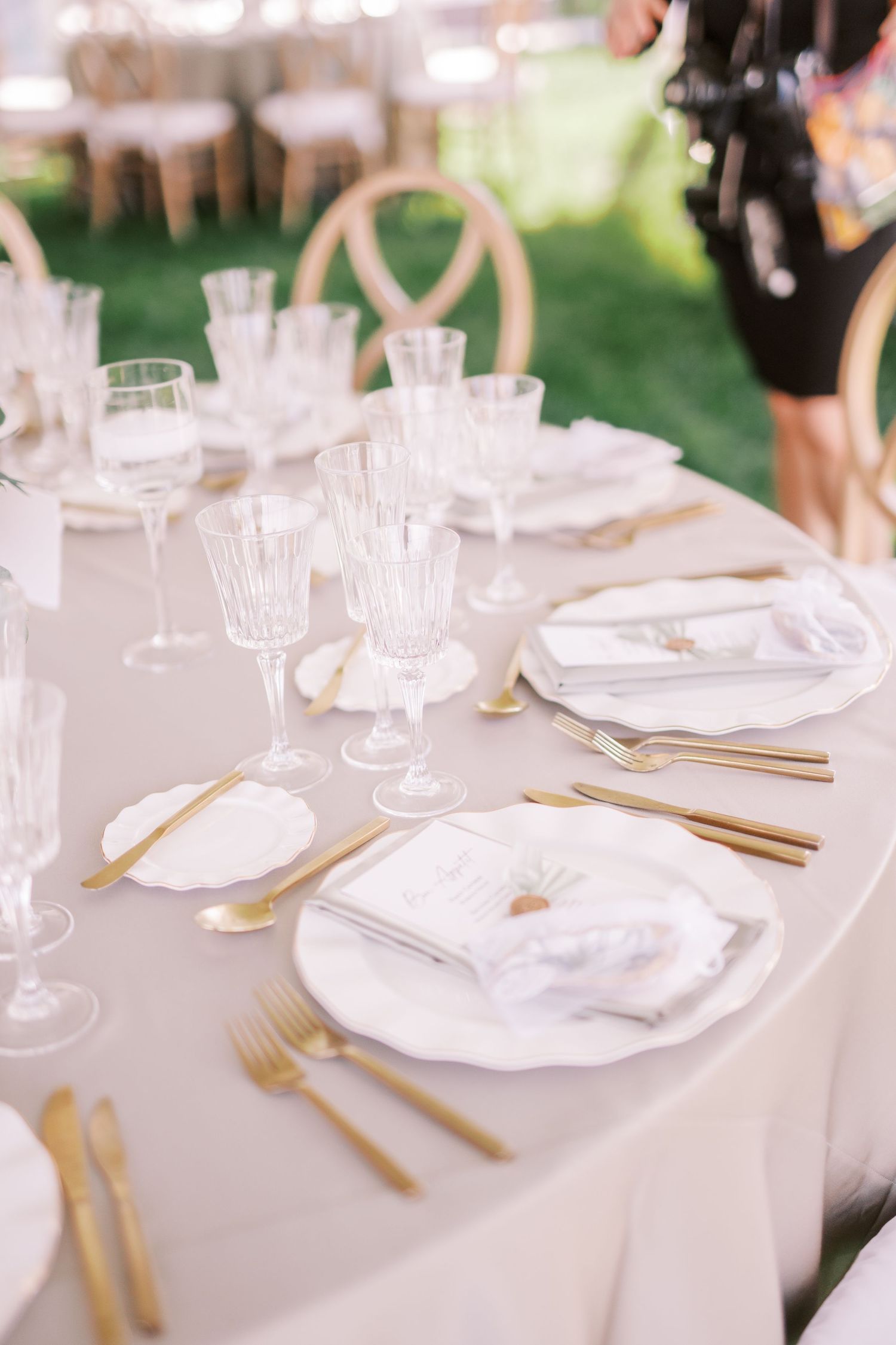 Elegant wedding table setting with white linens, gold cutlery, crystal glasses and white plates on an outdoor table.