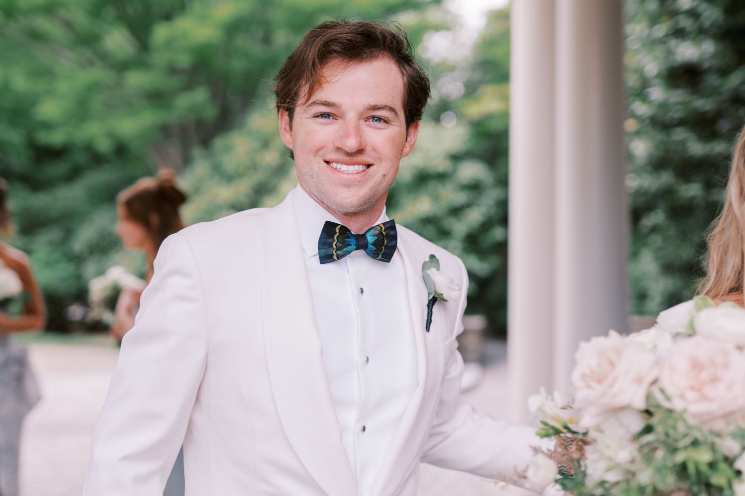 A traditional wedding party poses under white columns in an elegant outdoor garden setting.