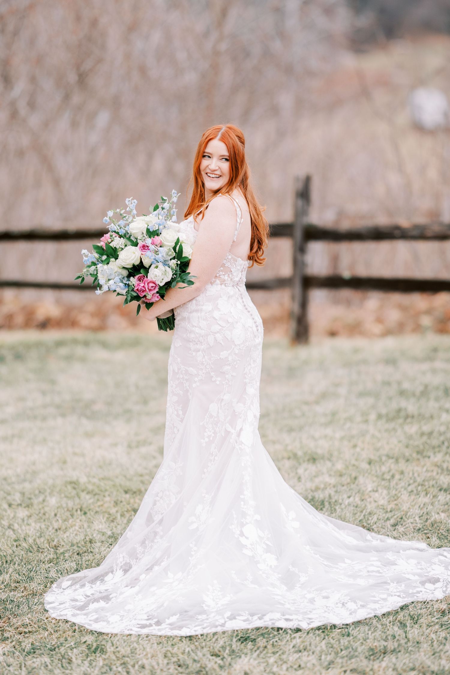 A bride in a lace wedding gown holds a pastel bouquet while posing in front of a rustic fence.