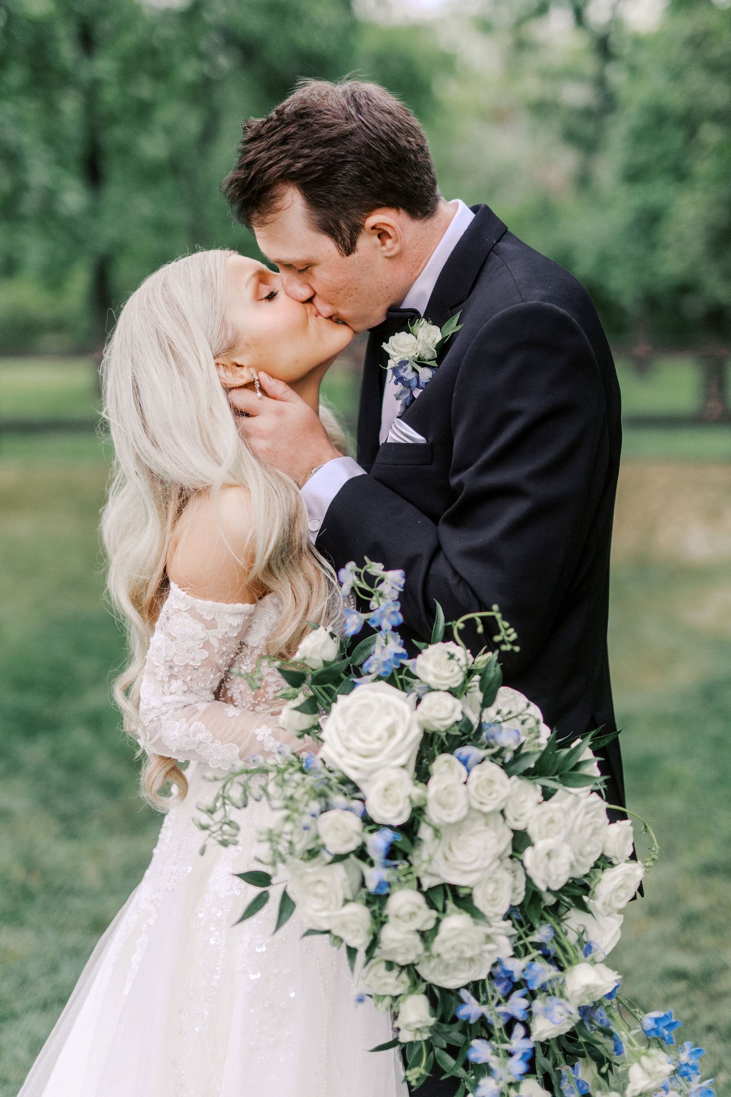 A romantic wedding moment captured outdoors with a stunning white bouquet featuring roses and blue accents.