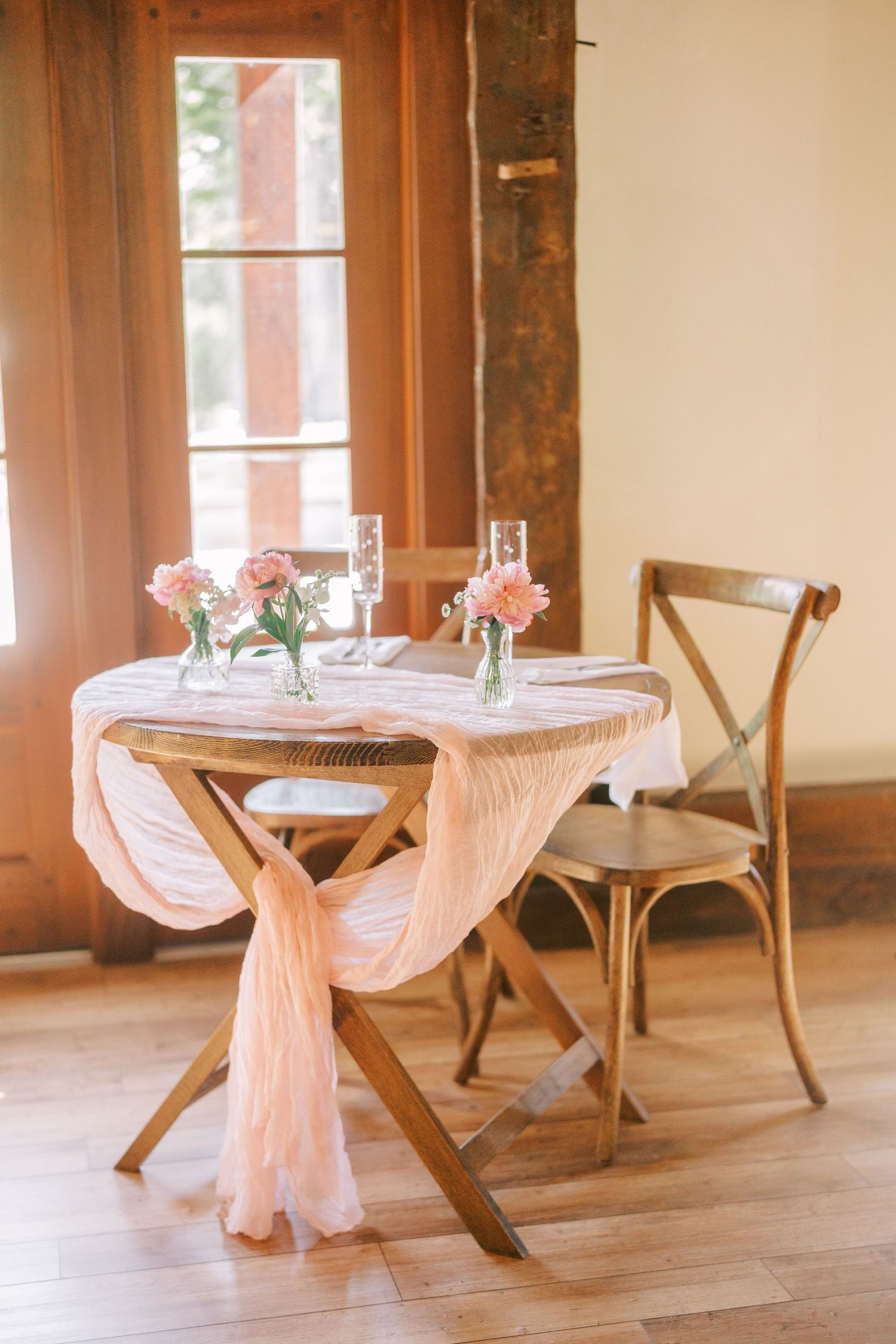 Rustic wooden reception tables with blush pink draping and place settings in a warm-lit venue with large windows.