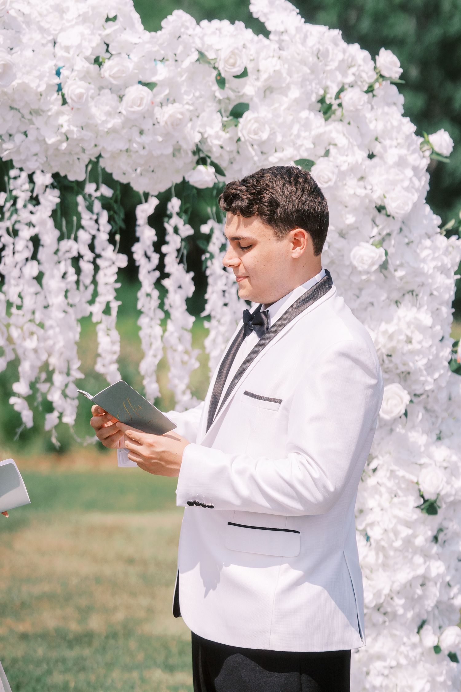 Someone in a white dinner jacket reads from a document under a cascading white floral wedding arch decoration.