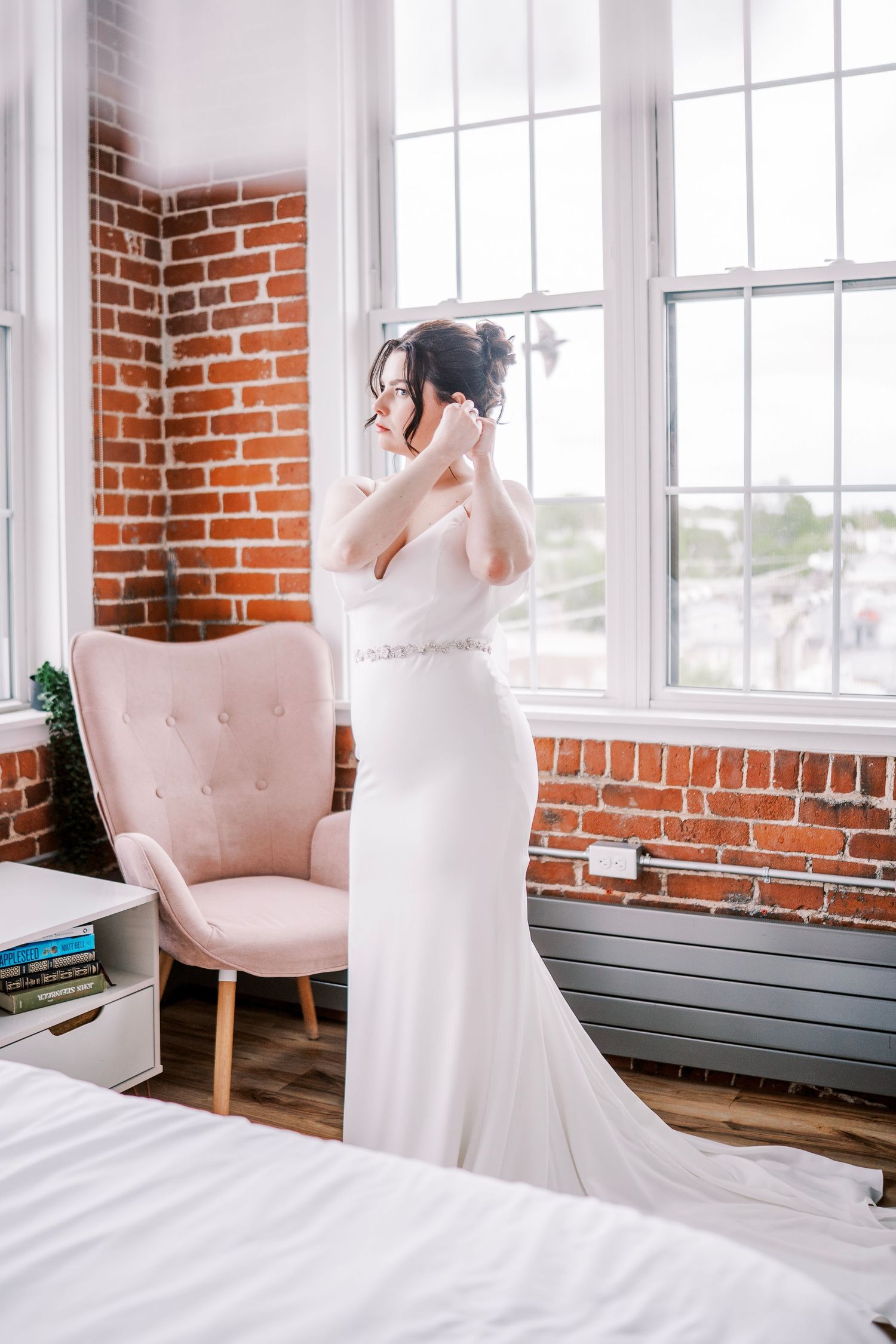 A bride in a fitted white wedding gown stands near a pink chair in an industrial-style room with brick walls.