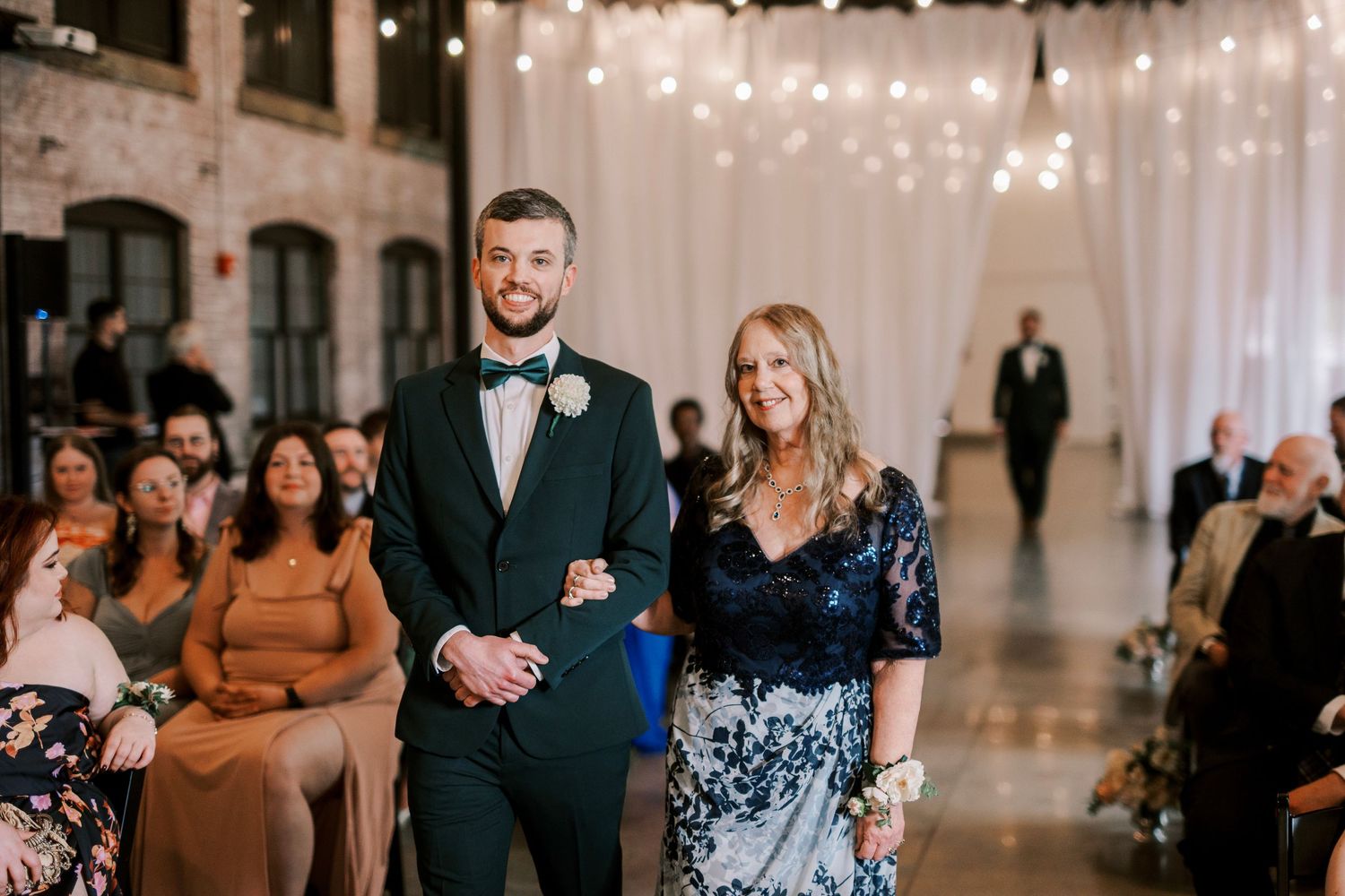 A couple walks down the aisle at an indoor wedding ceremony with string lights and white draping.