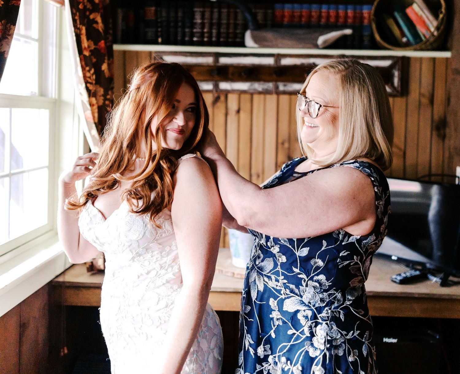 A person in a blue floral dress helps adjust a white lace gown on someone by a window in a rustic room setting.