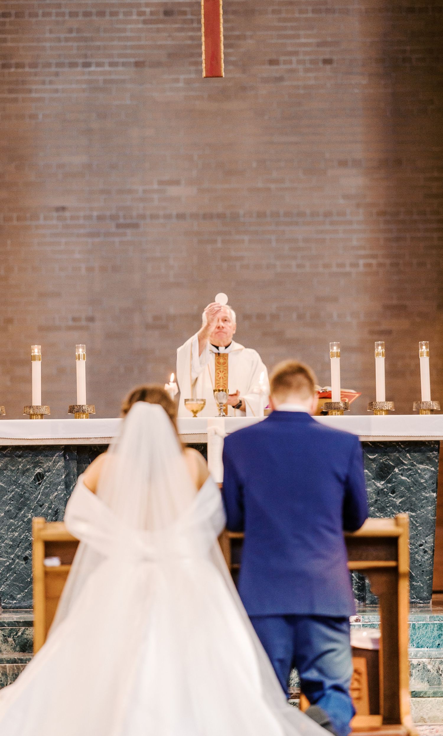 Traditional church wedding ceremony with white candles and marble altar as couple kneels before priest at the altar.