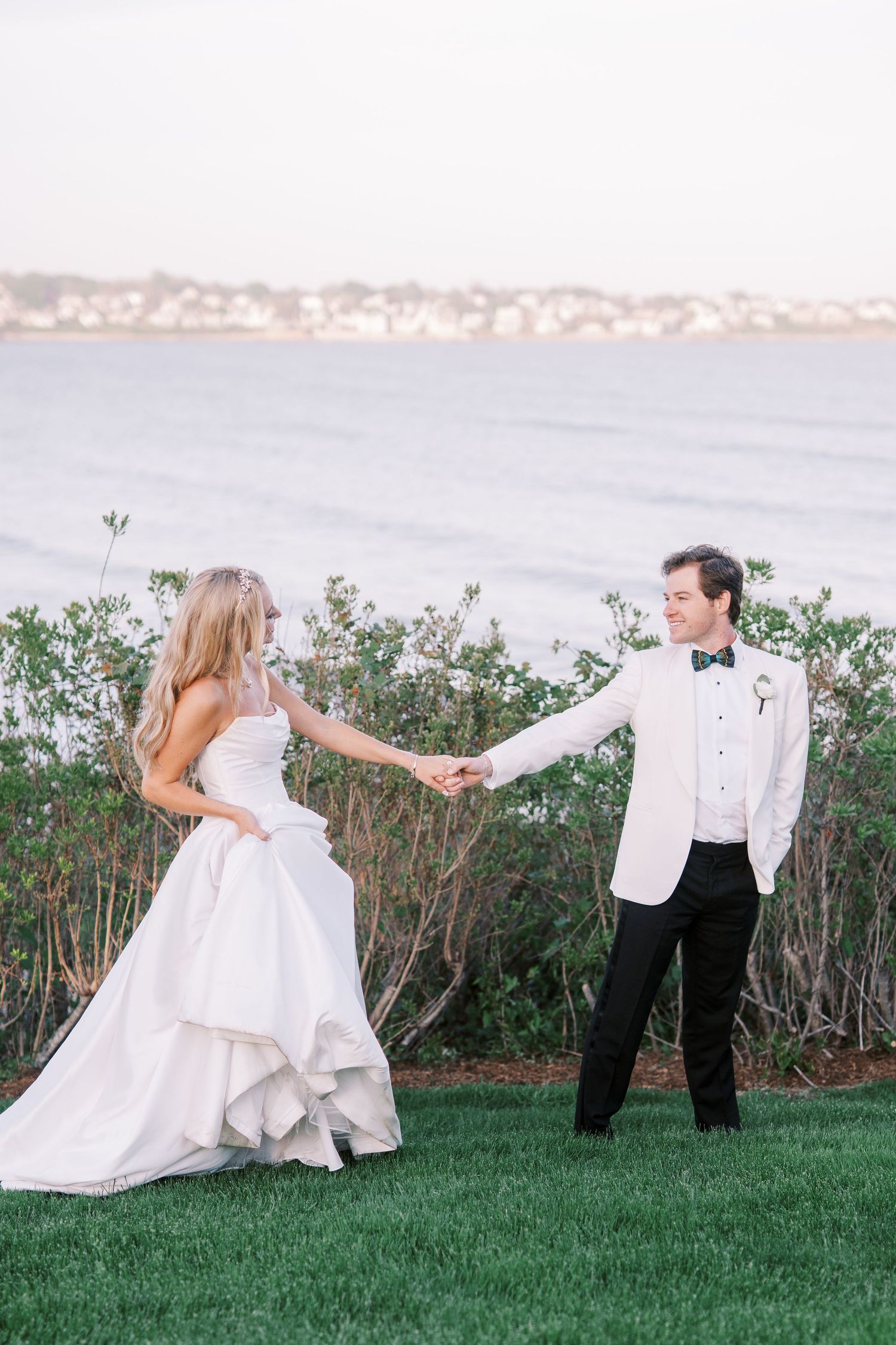 A couple in wedding attire dances playfully on a grassy lawn with ocean and hills visible in the background.