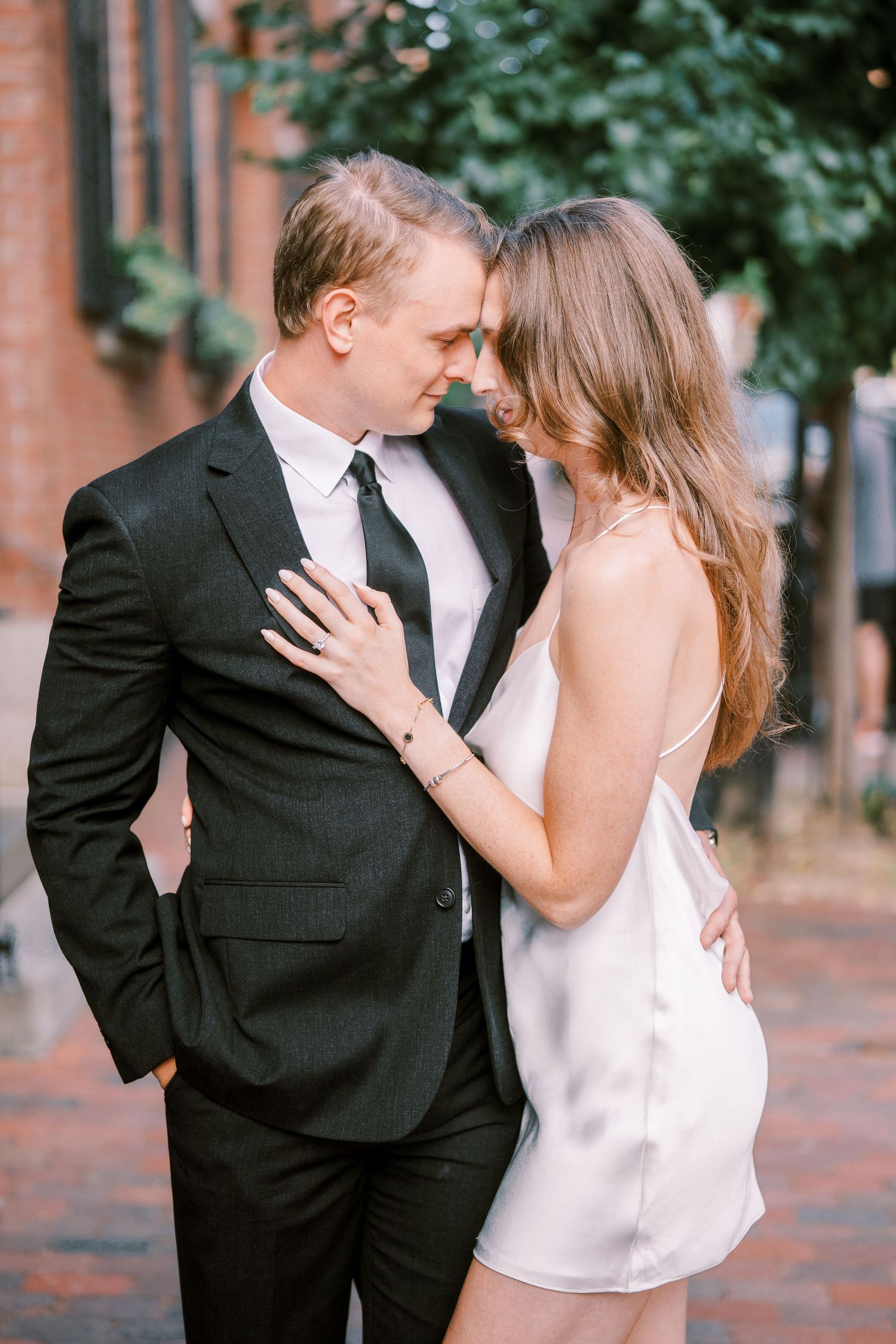 Romantic couple embraces in formal attire on a cobblestone street with brick buildings and greenery in the background.