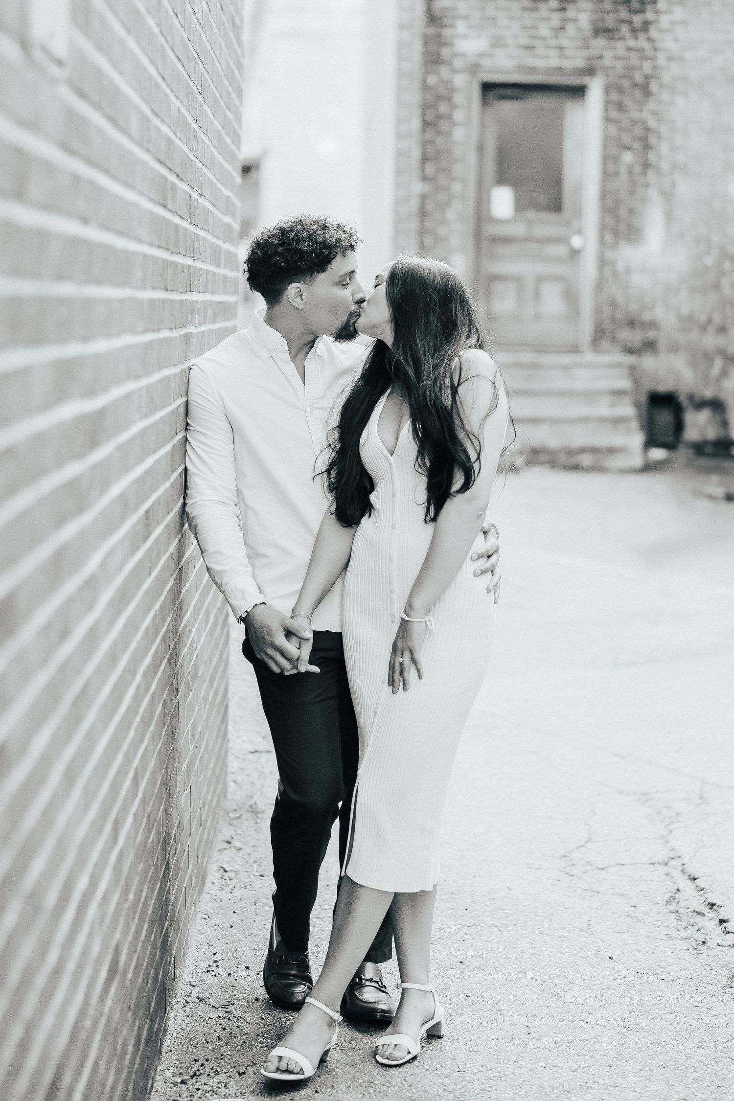 A couple shares intimate moments against a white brick wall in black and white photography.
