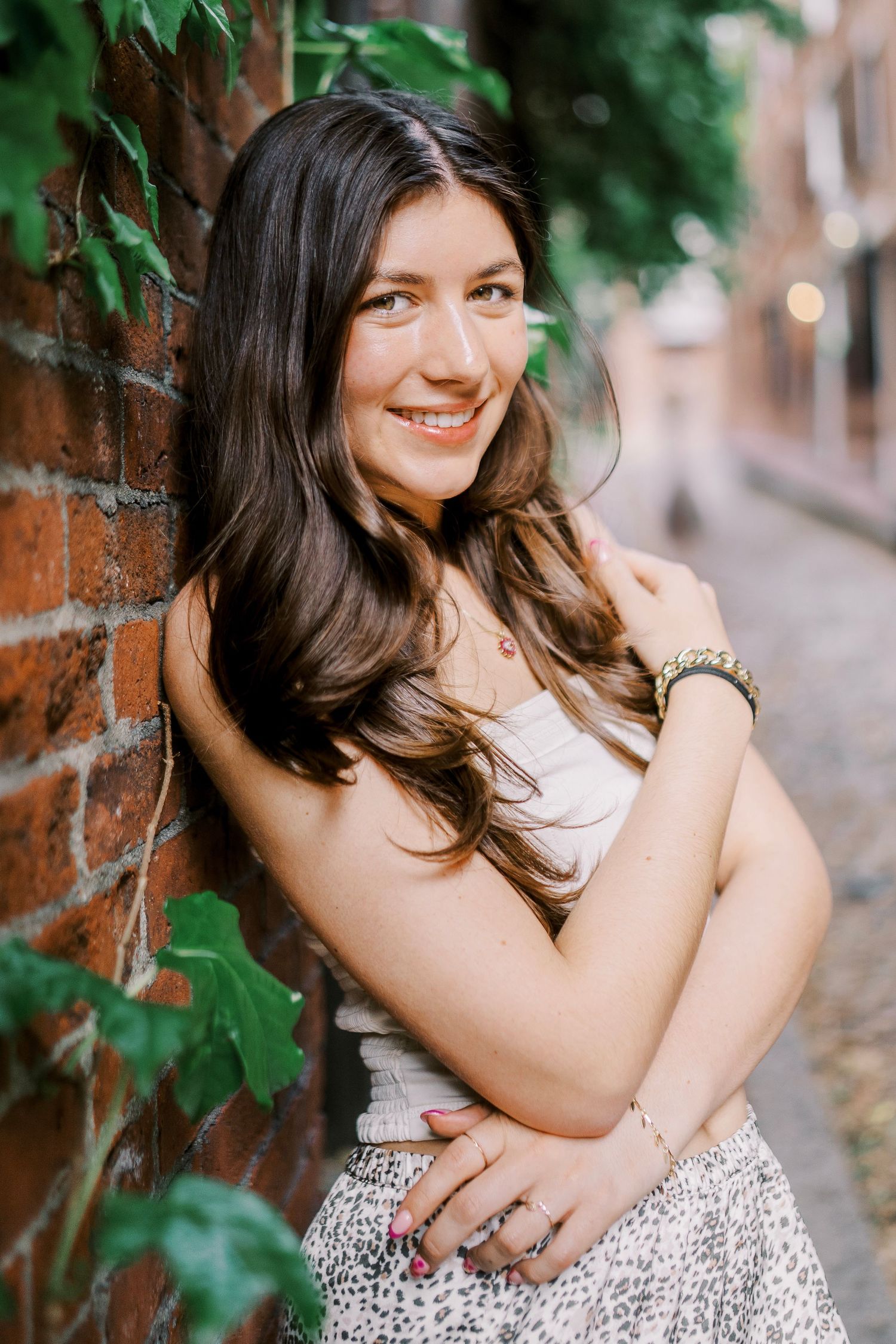 Portrait of a person in a white top and patterned skirt leaning against a brick wall with green foliage.