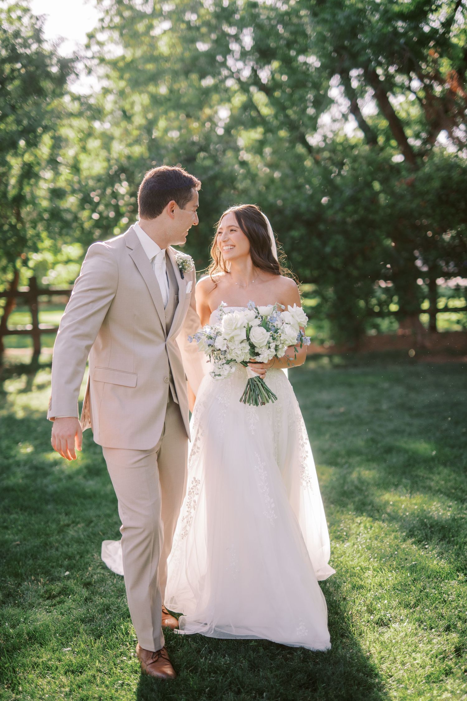 A couple walks together on lush green lawn, sharing a joyful moment in elegant wedding attire and holding white bouquet.
