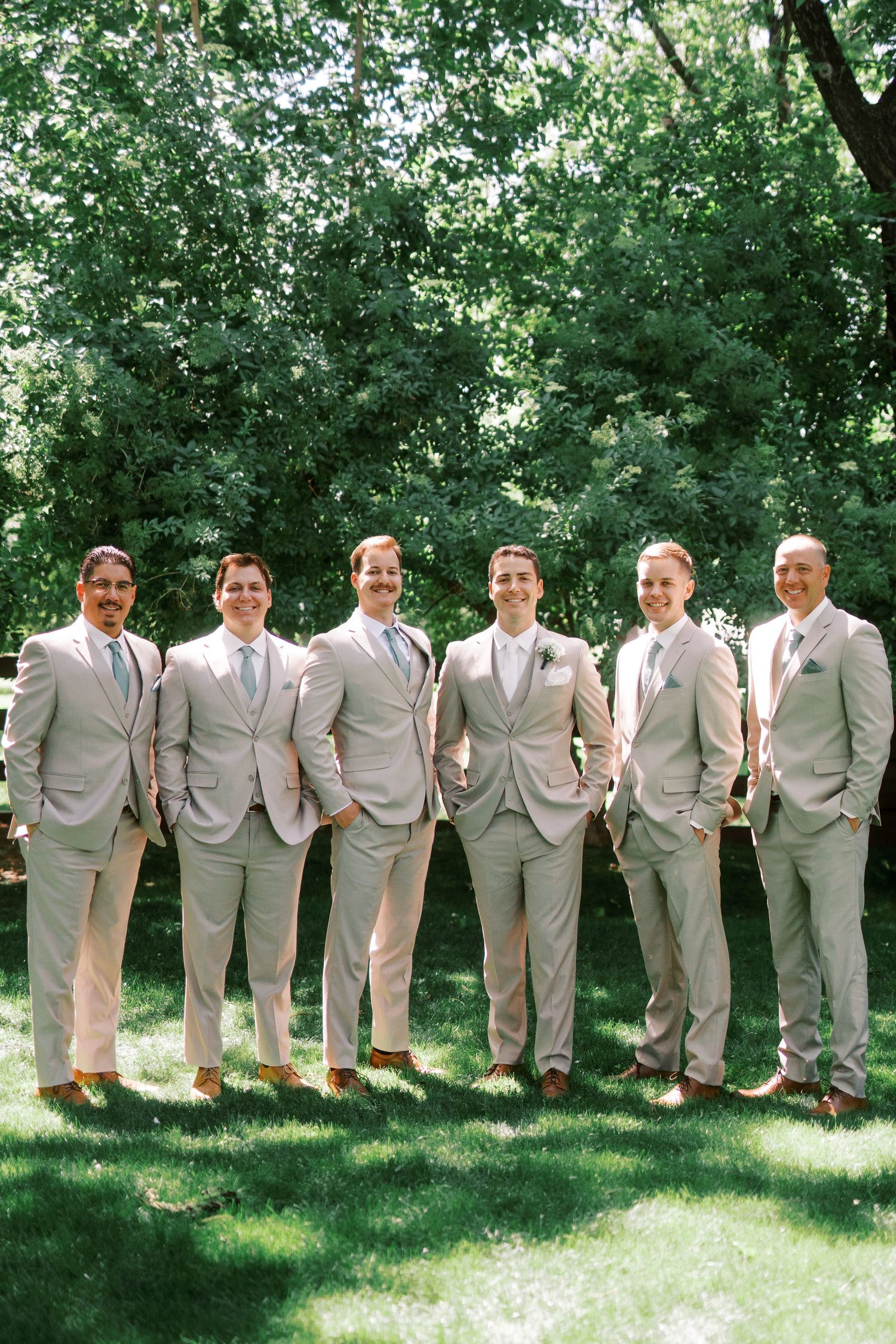 Groomsmen in light gray suits stand side by side on grass with trees creating a natural backdrop.