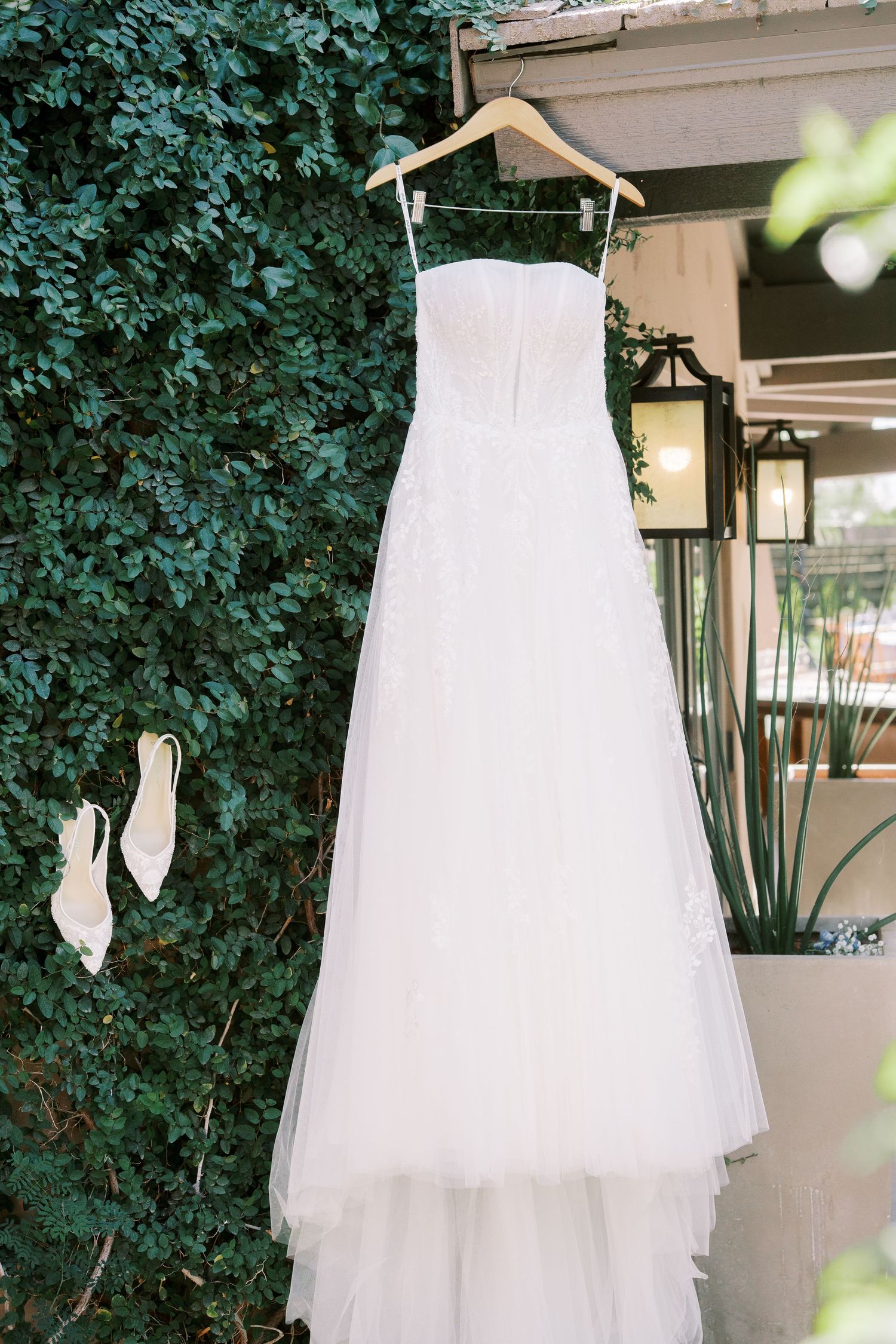 A white wedding dress with tulle skirt hangs on a wooden hanger outdoors with matching white shoes against green foliage.