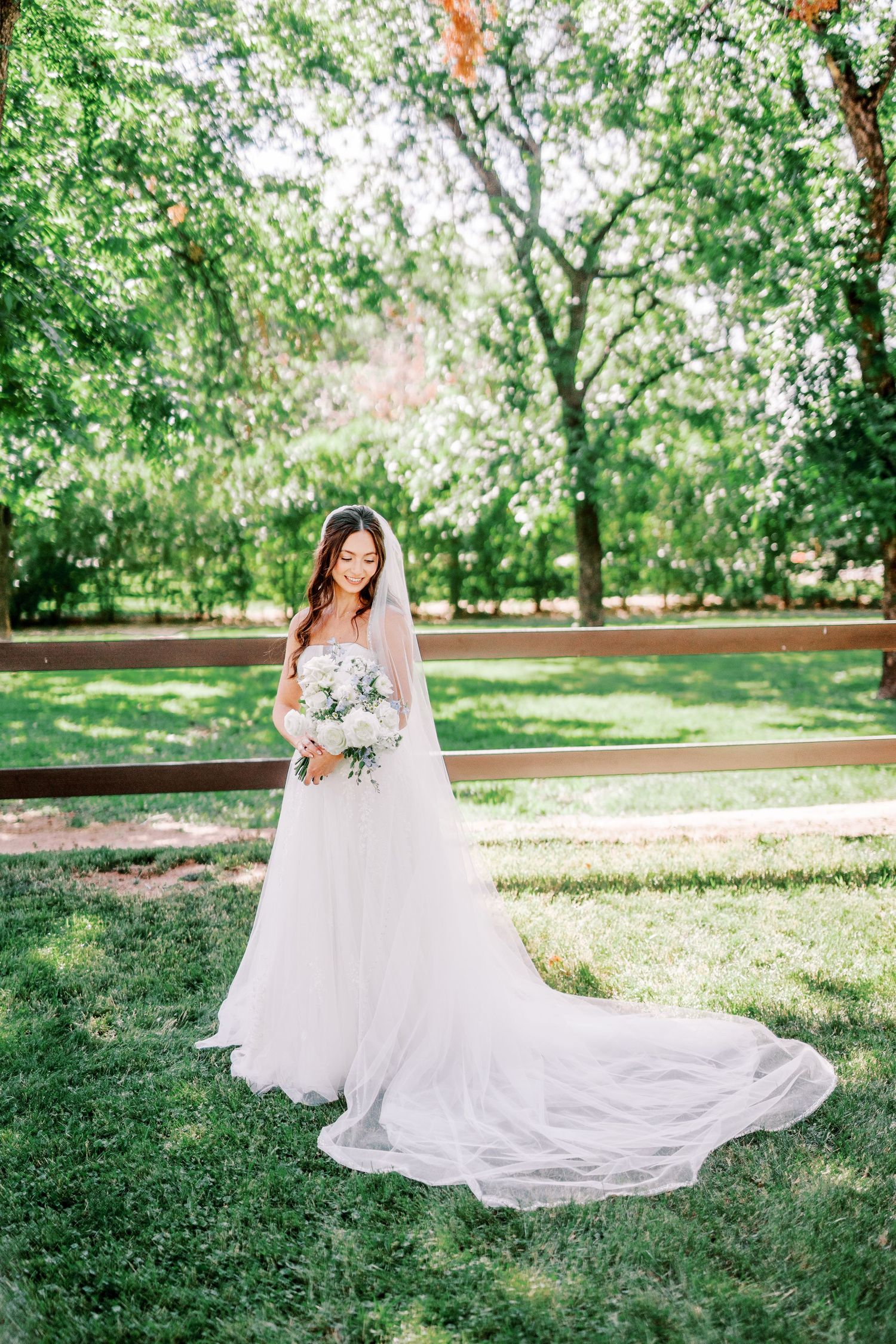 A bride in a white lace wedding gown with long train poses on a sunny day in a lush green garden setting.