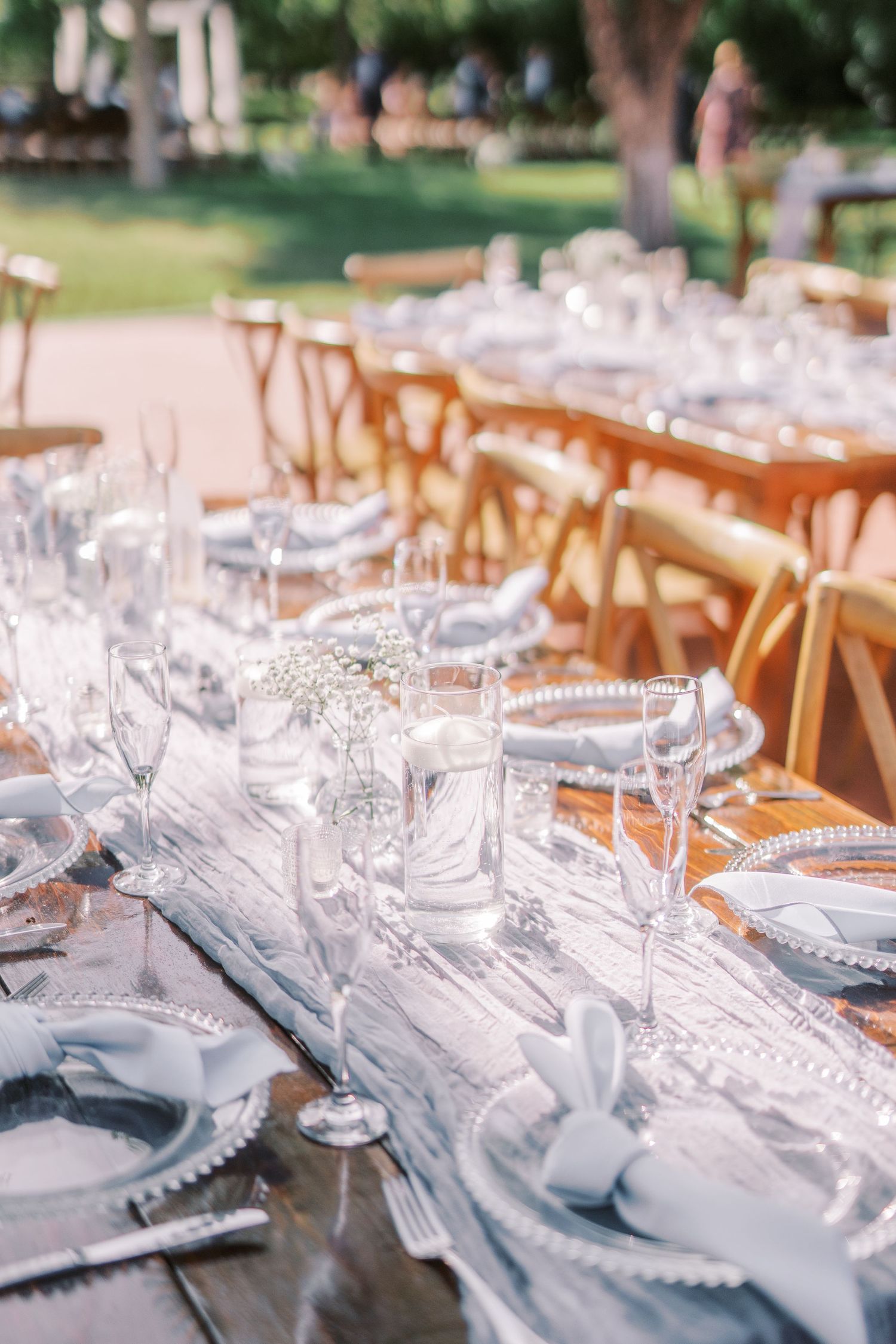 Elegant outdoor wedding reception table setting with rustic wooden chairs and delicate glassware on a white tablecloth.