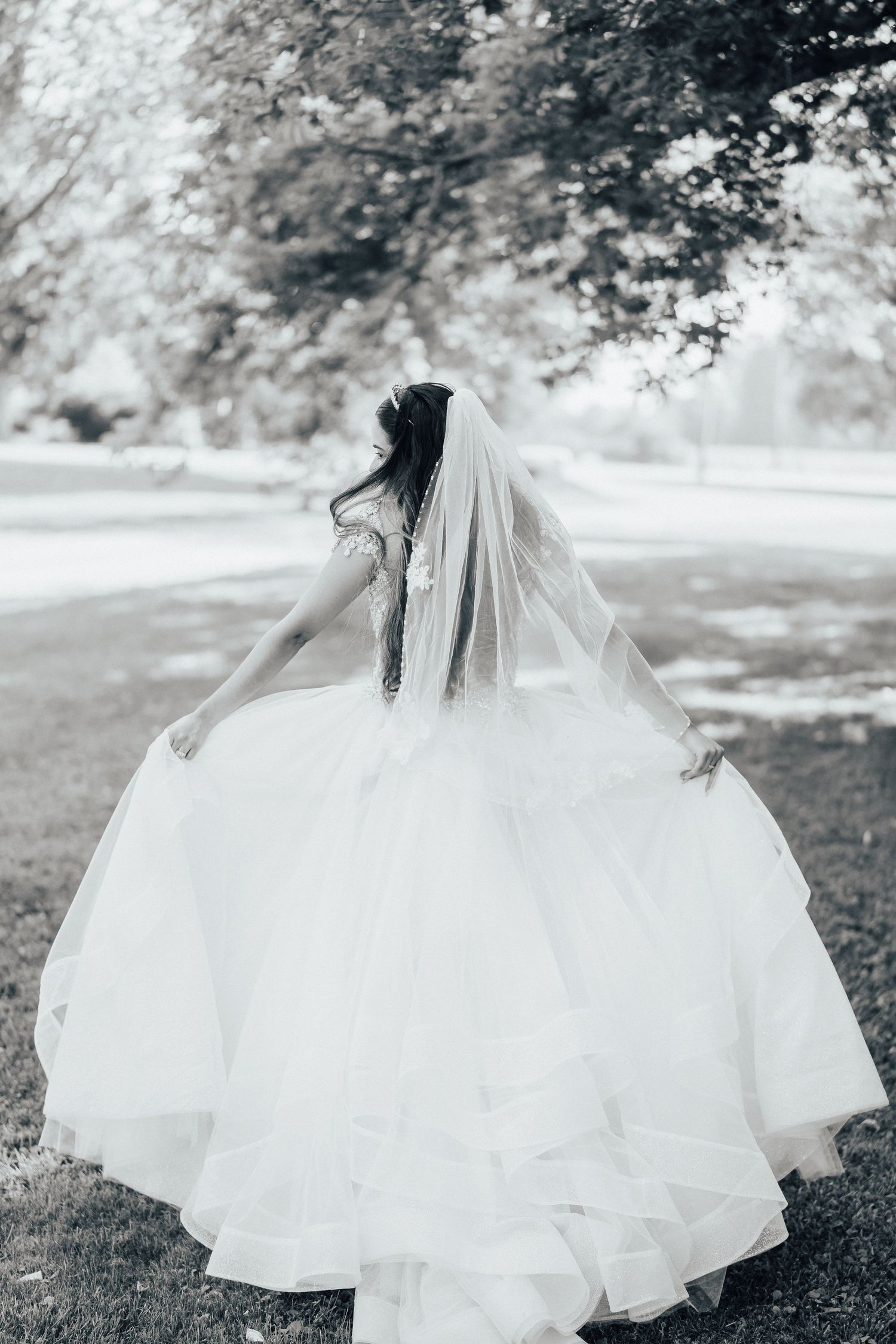 A bride in a flowing white wedding gown twirls in a garden setting, photographed in black and white.