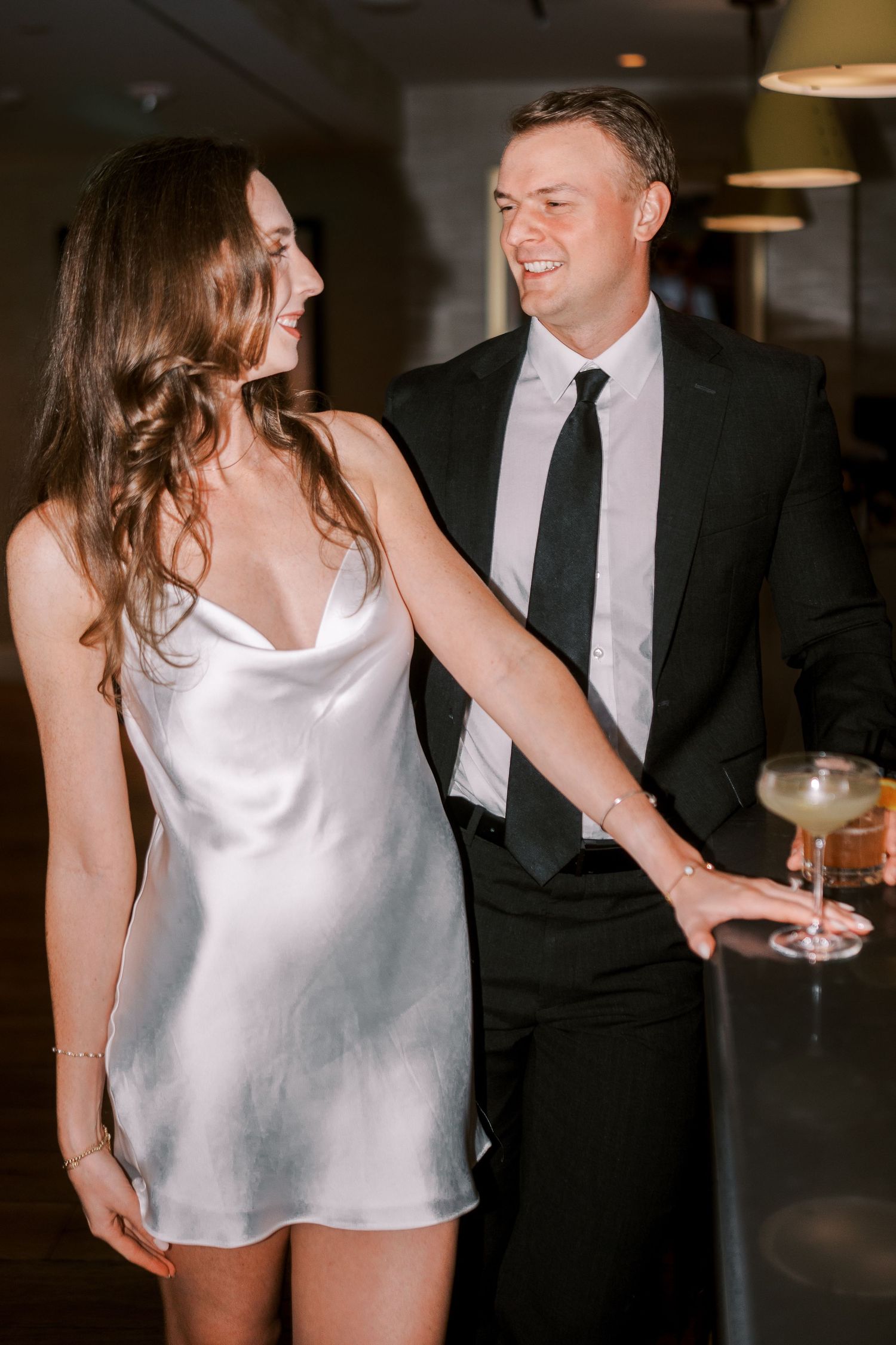 Two people in evening attire smile and laugh together at an indoor event while holding cocktail glasses.