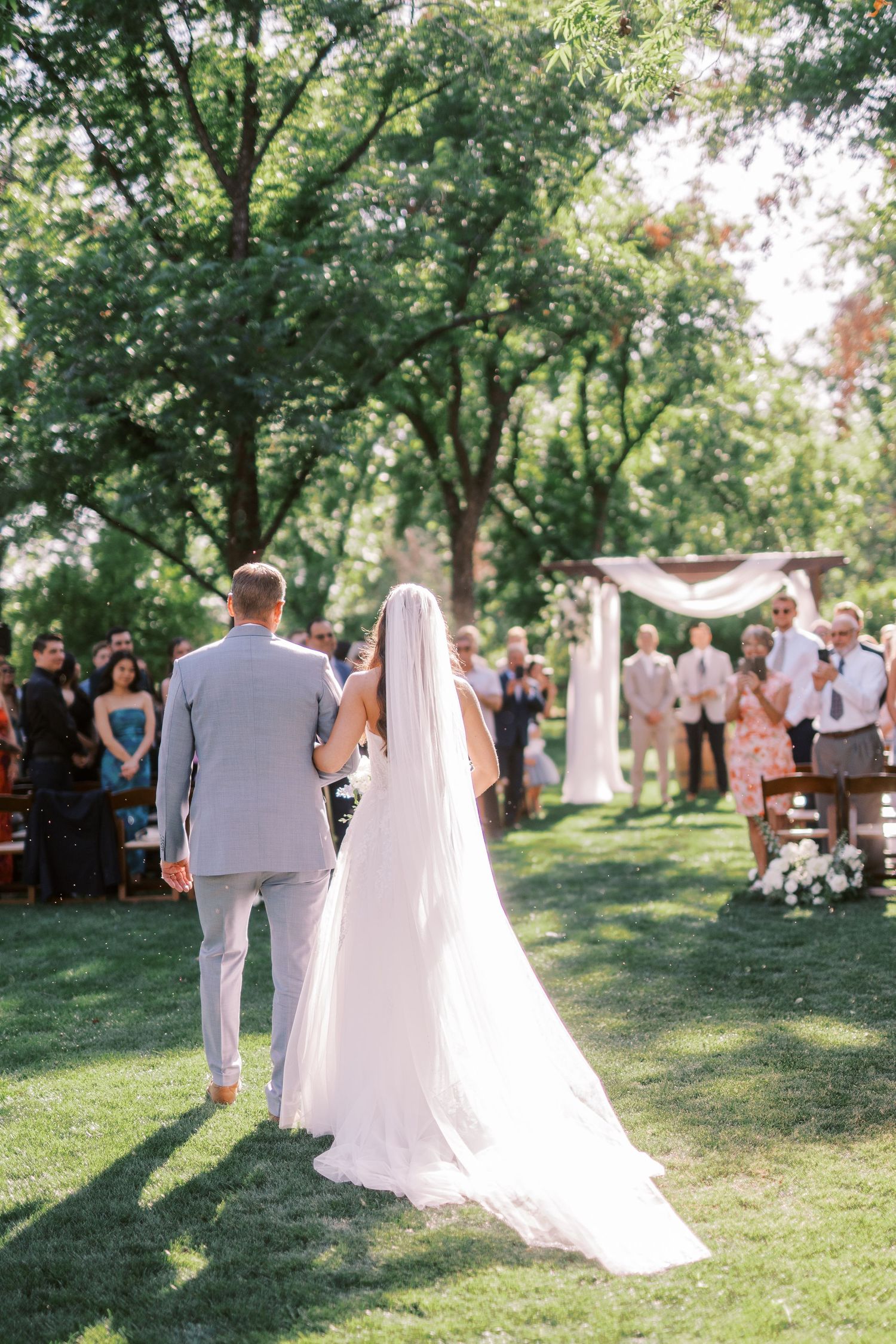 Couple walking down a tree-lined outdoor ceremony aisle on a sunny summer day with guests and white floral decorations.