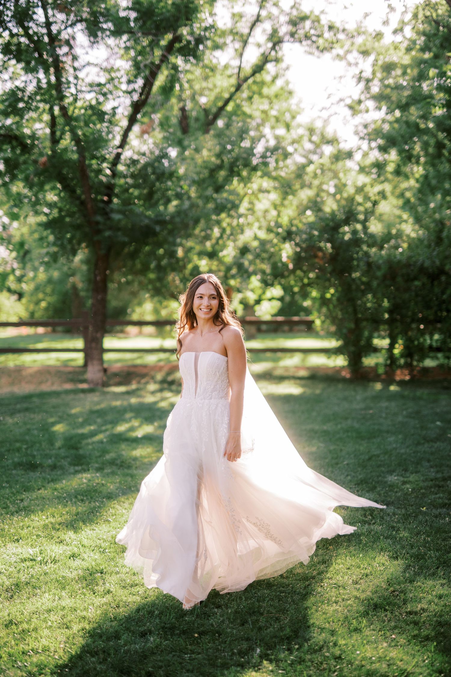 A bride in a flowing white gown gracefully twirls on a sunlit lawn surrounded by mature trees.