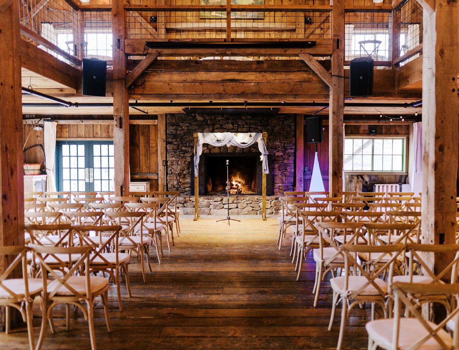 Rustic barn wedding venue with wooden cross-back chairs, stone fireplace and exposed timber beams in warm lighting.