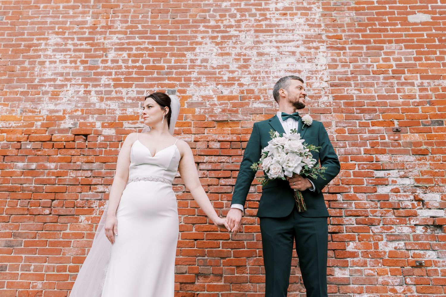 Wedding couple poses against rustic brick wall holding hands and bouquet.