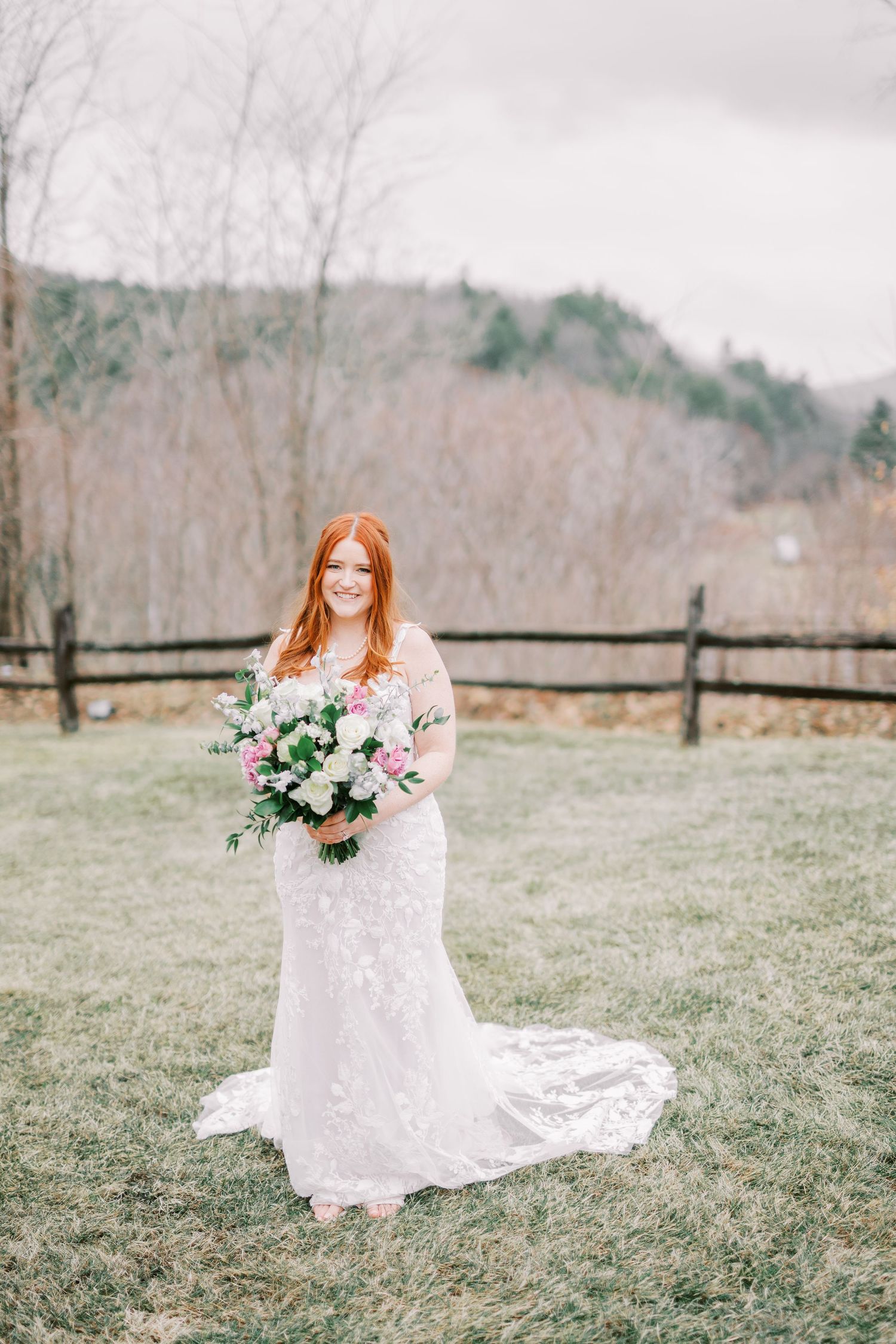 Bride in lace wedding dress holds pink and green bouquet while standing in field with rustic fence and winter trees.
