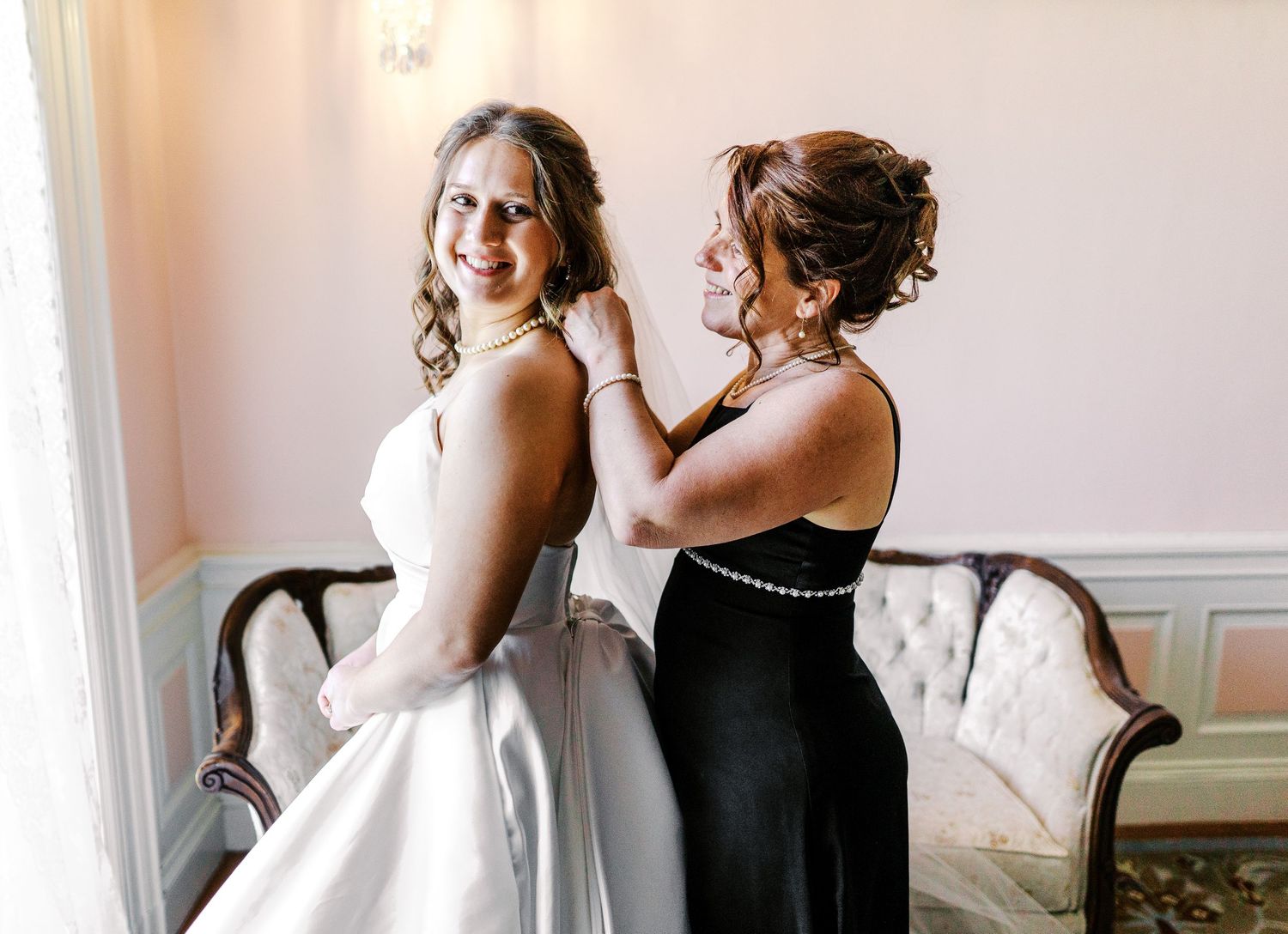 Someone helps adjust a classic white wedding gown against pink walls in an elegant room with tufted furniture.