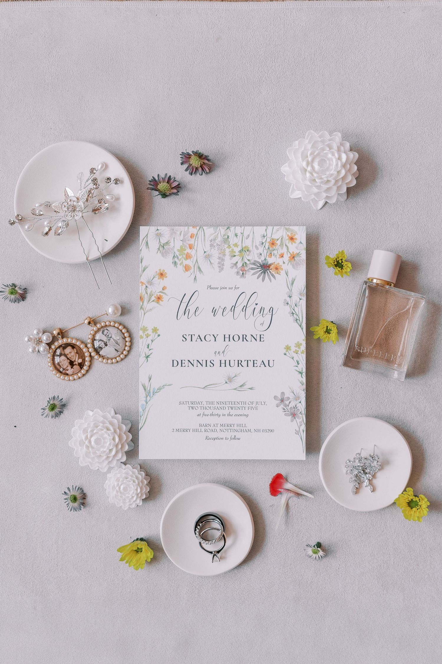 Wedding details flatlay featuring invitations, rings, perfume, and delicate white decorative elements on a light gray surface.
