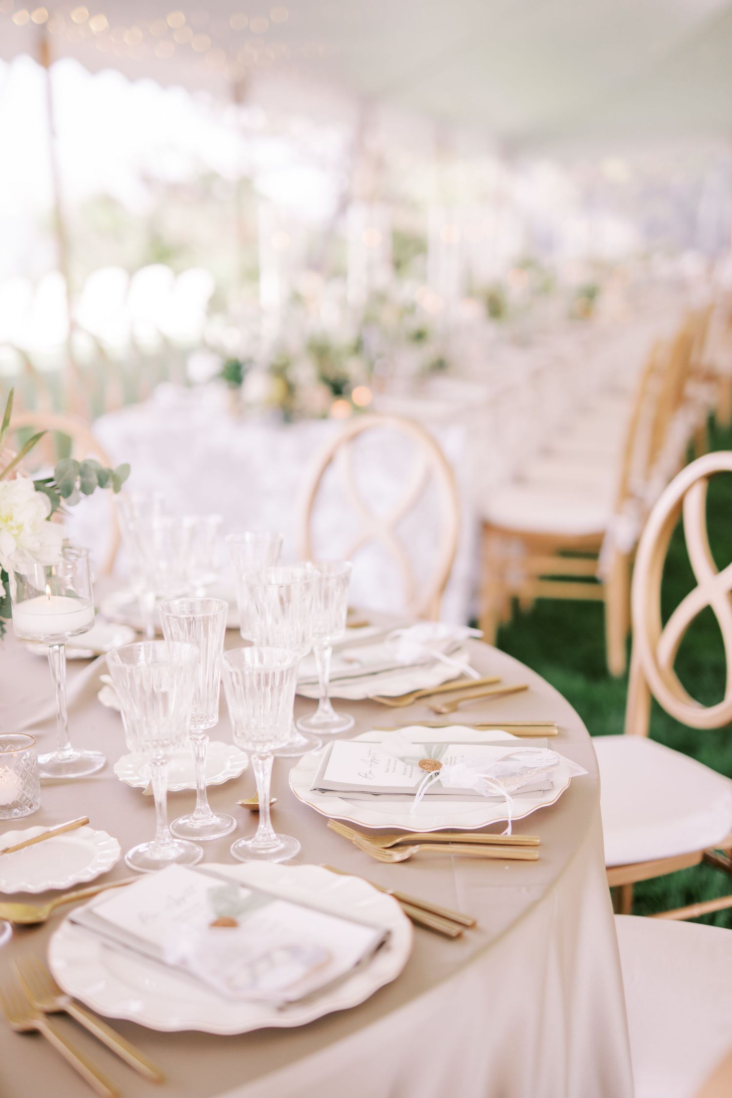 Elegant wedding table setting with crystal glasses, white plates and gold accents on beige linens at an outdoor reception.