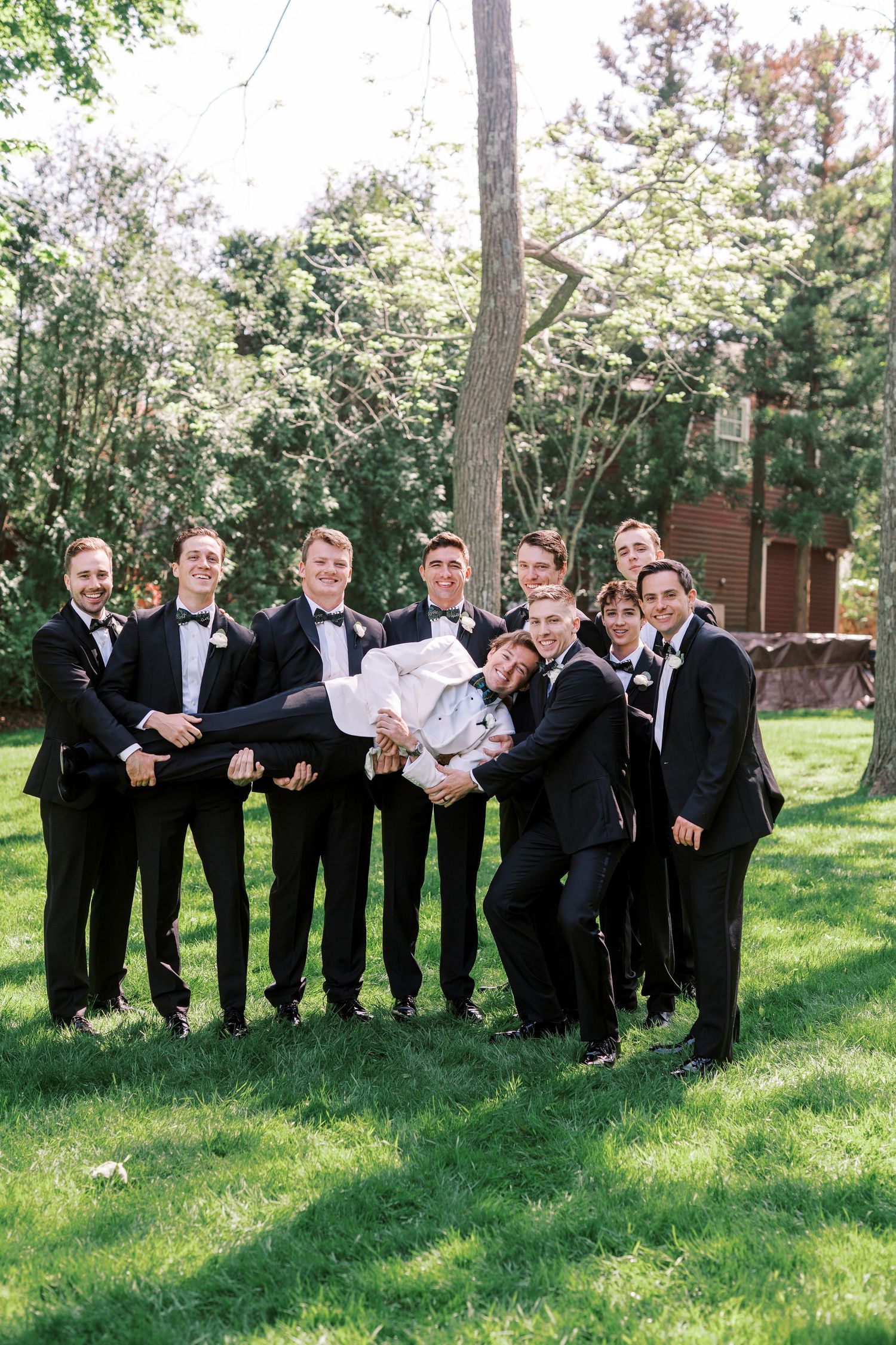 A group wearing black tuxedos and bowties pose together on a sunny lawn with trees and greenery in the background.