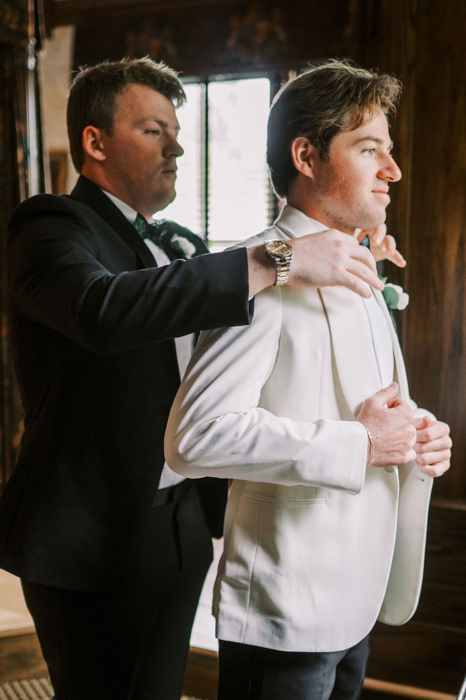Best man helps groom adjust his white tuxedo jacket while getting ready for wedding ceremony in rustic venue.