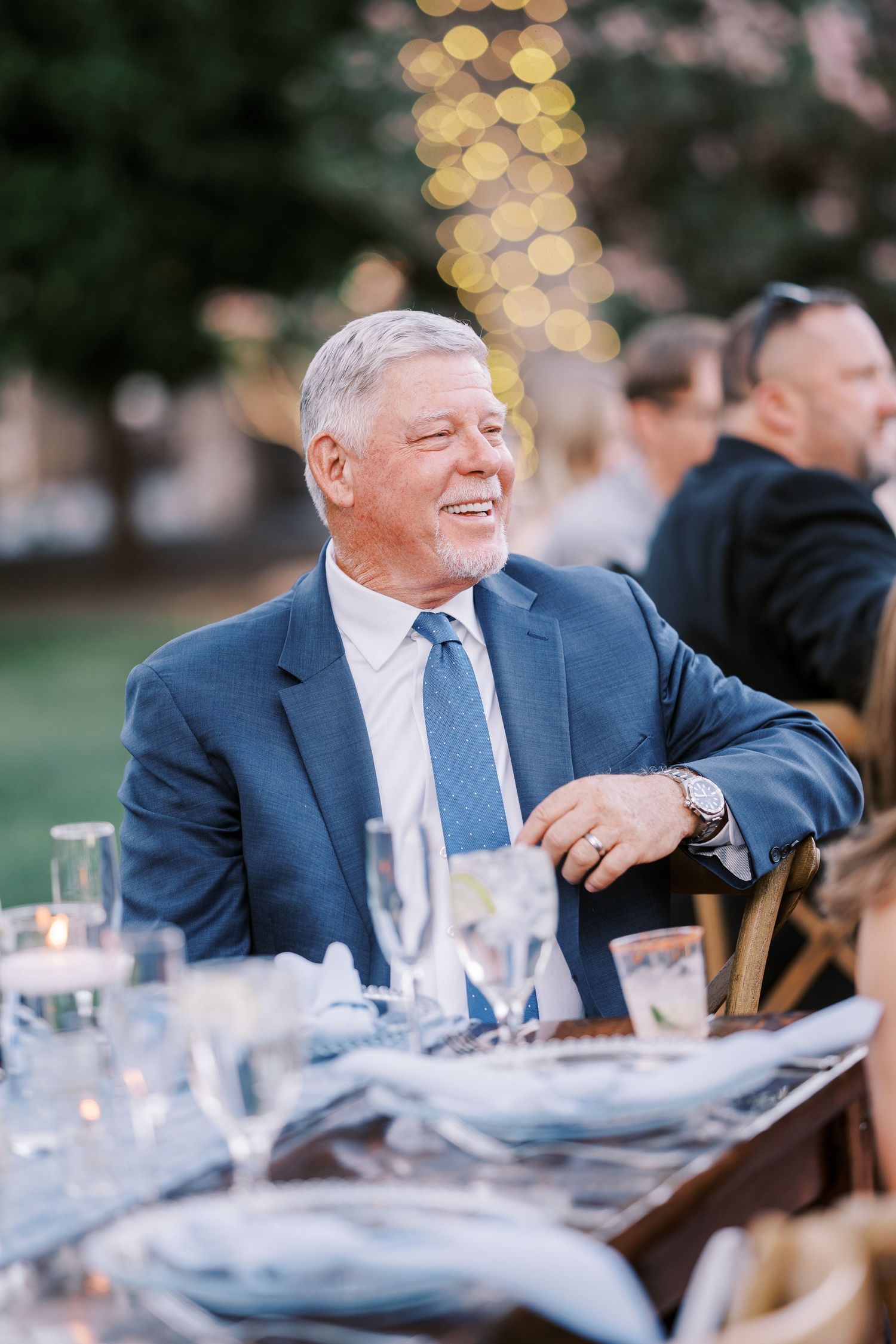 A guest in a navy suit sits at an elegant outdoor dinner table with bokeh lights in the background.