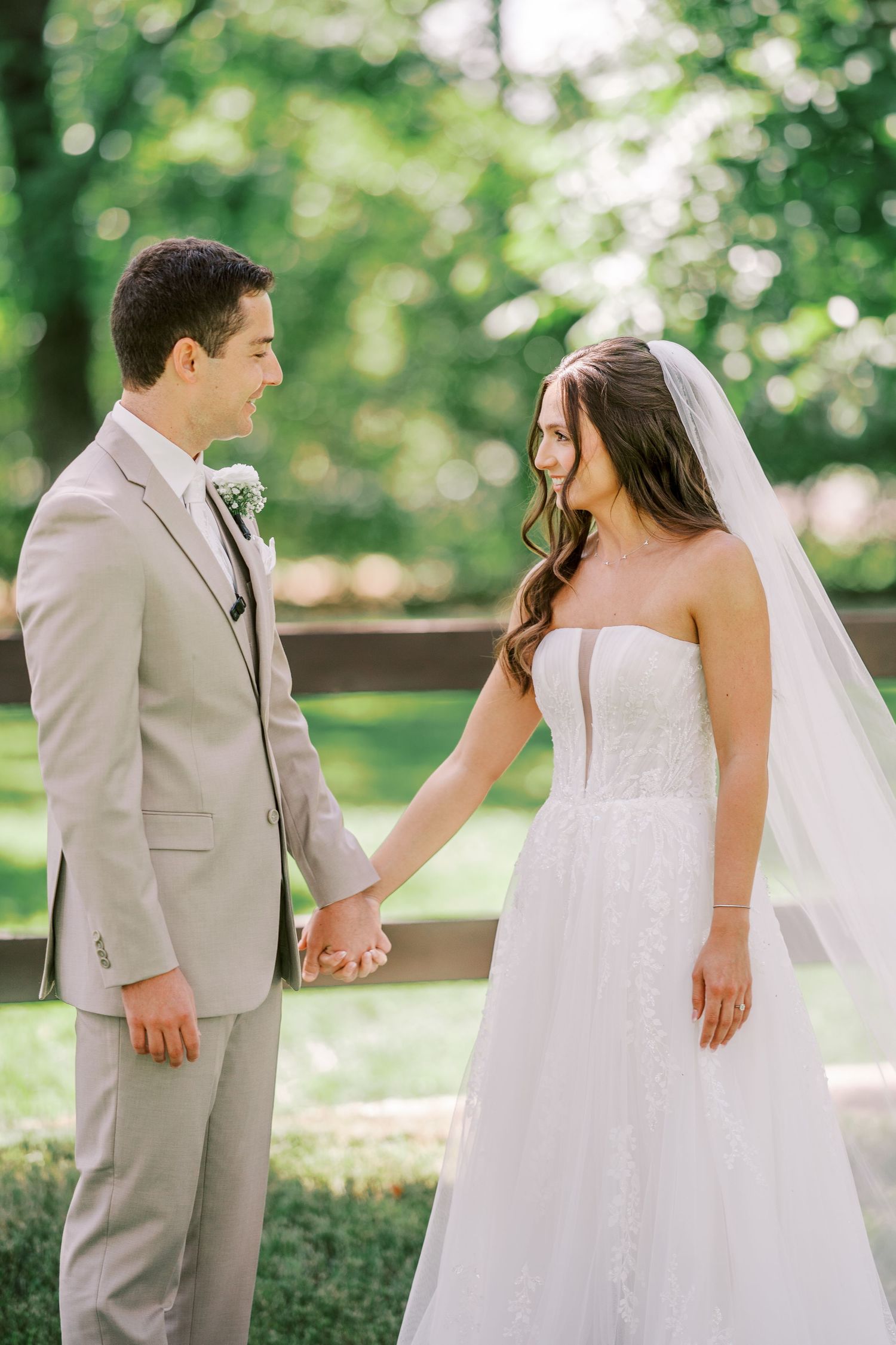 A couple in wedding attire holds hands while standing in a lush garden with soft natural lighting.