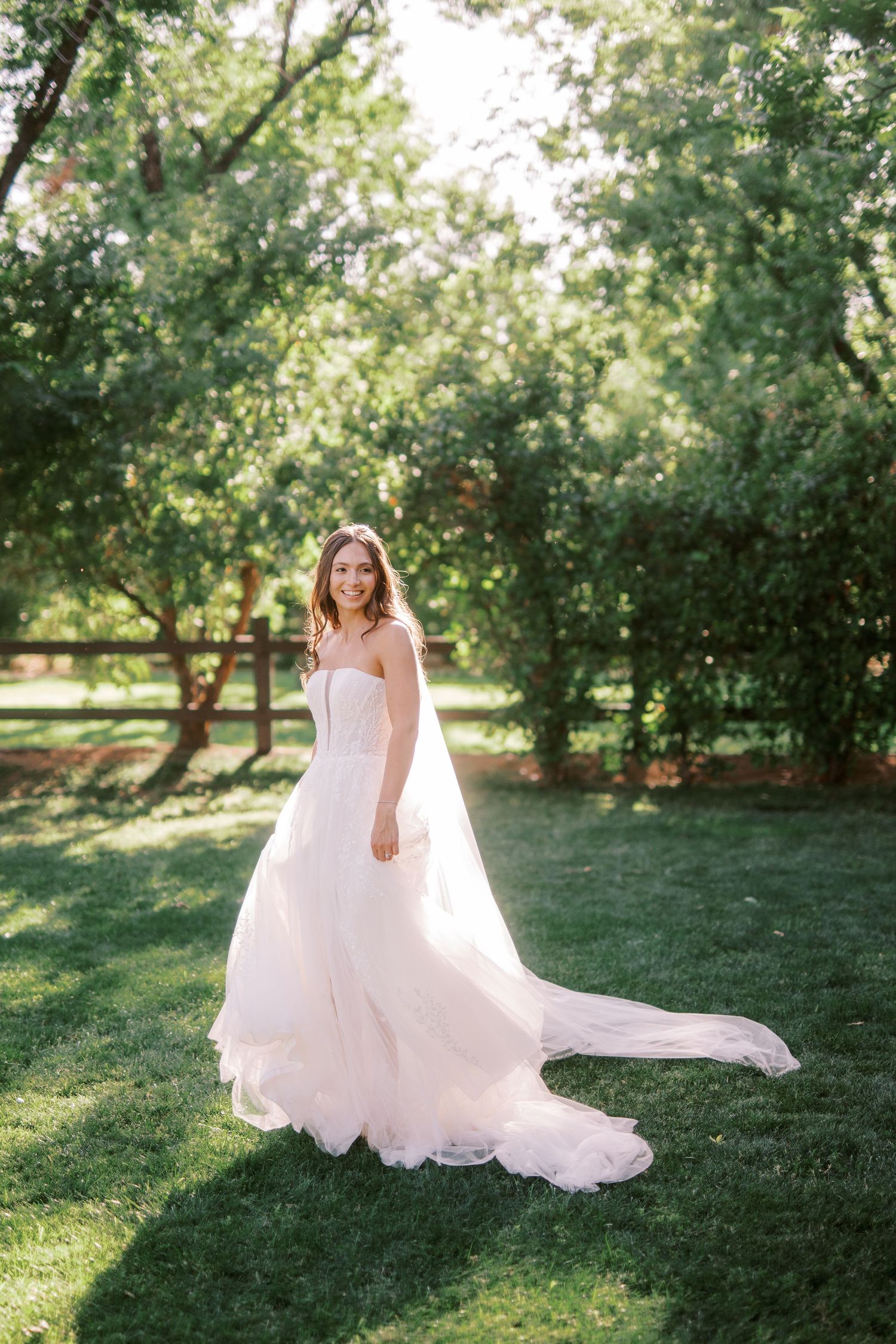 A beautiful wedding dress with flowing train photographed on lush green lawn with sunlit trees in background.